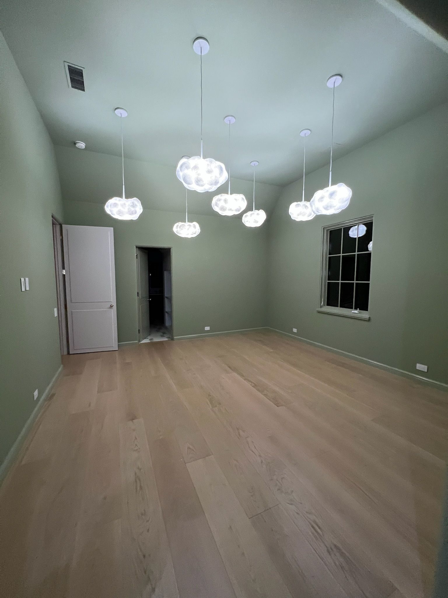 Light-colored hardwood floor in a room with light green walls and eight hanging, cloud-shaped lights. A door and window are present.