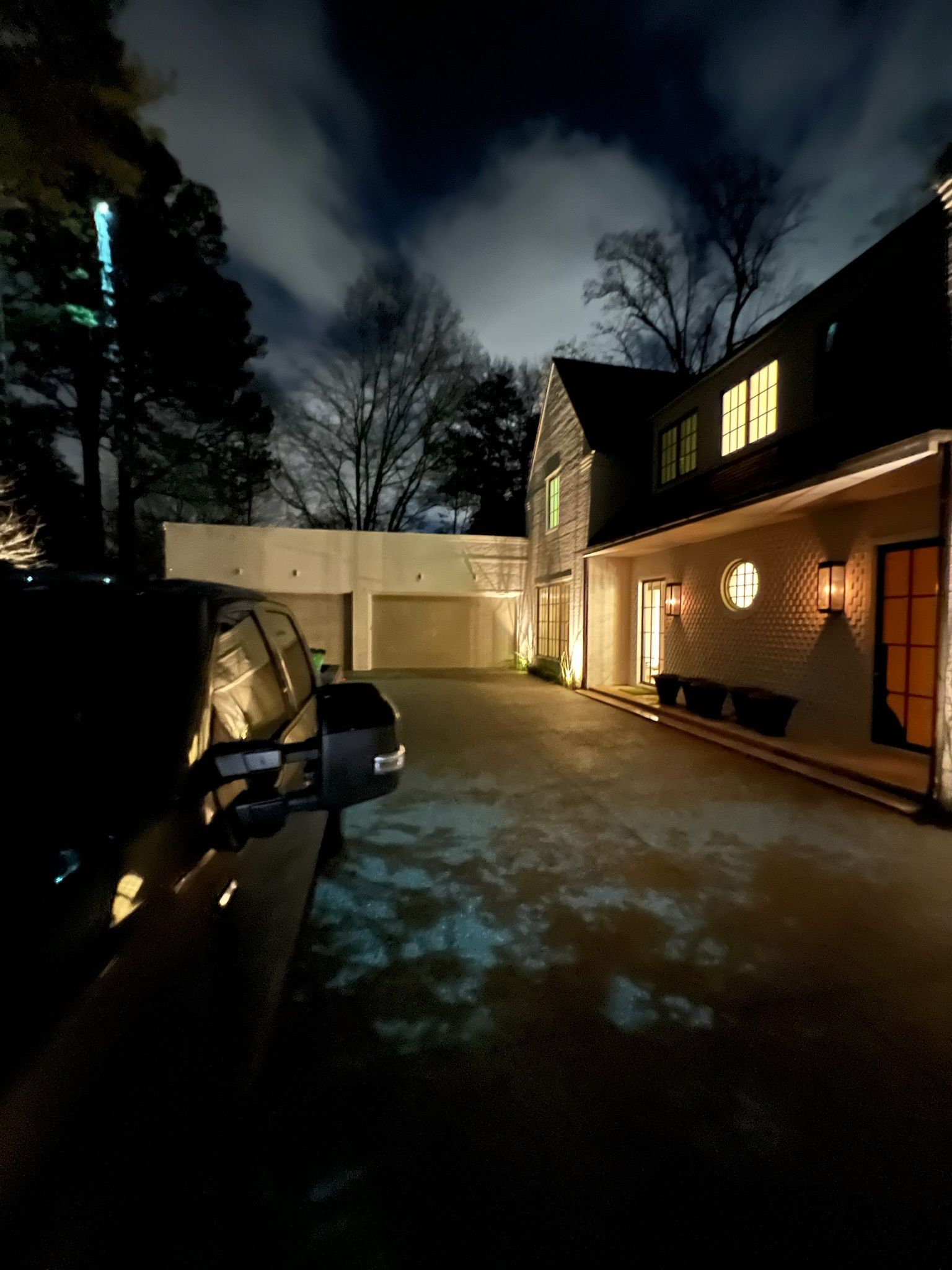 Night view of a house and driveway, lit by outdoor lights. A vehicle is parked in the foreground. Overcast sky.