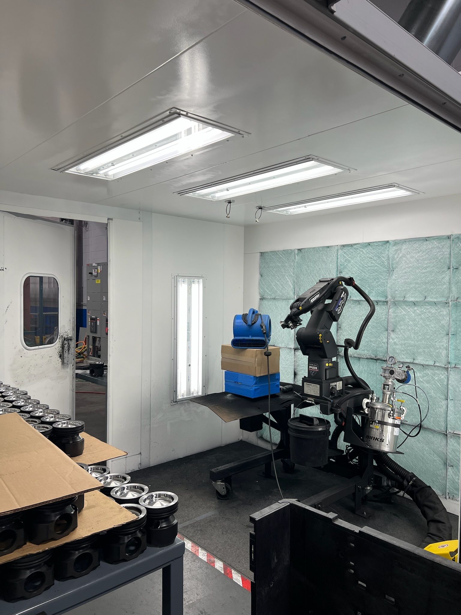 Robotic arm spray-painting parts inside a spray booth. Boxes and parts are present on a table and on the floor.