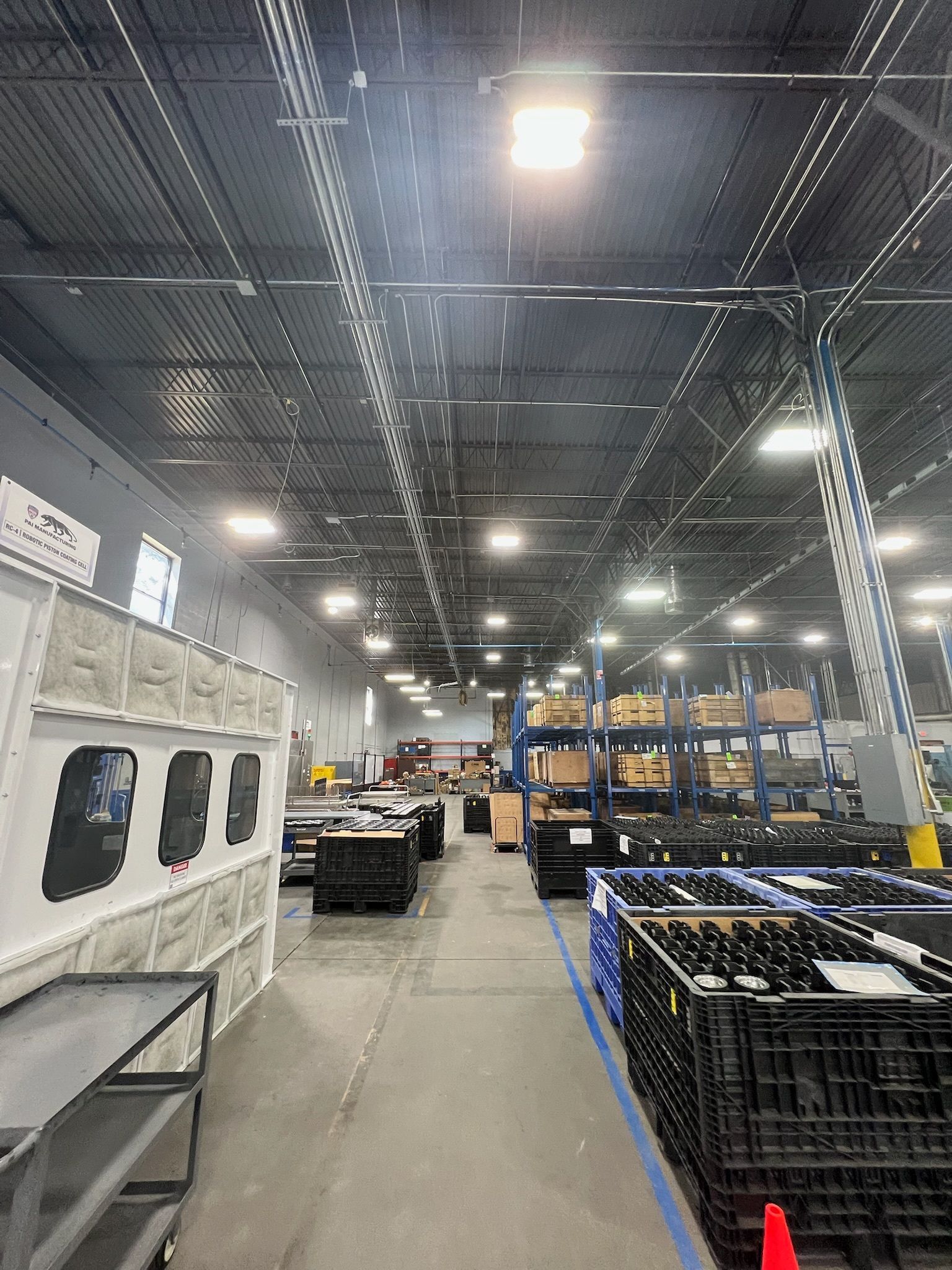 Warehouse interior with high ceiling, metal beams, and shelving with items. Black bins and a concrete wall are in the foreground.