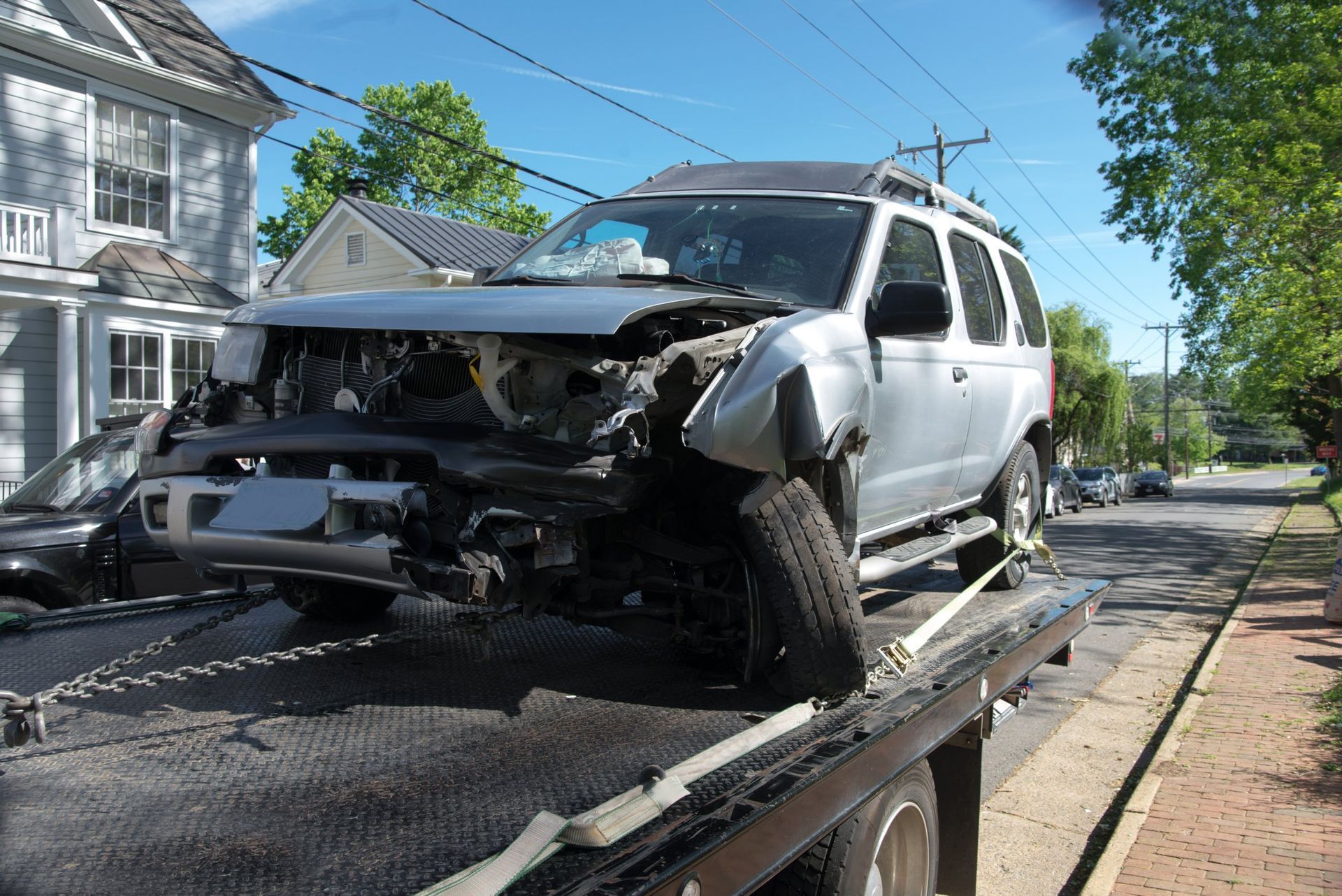Silver SUV severely damaged on a tow truck; street and houses in background.