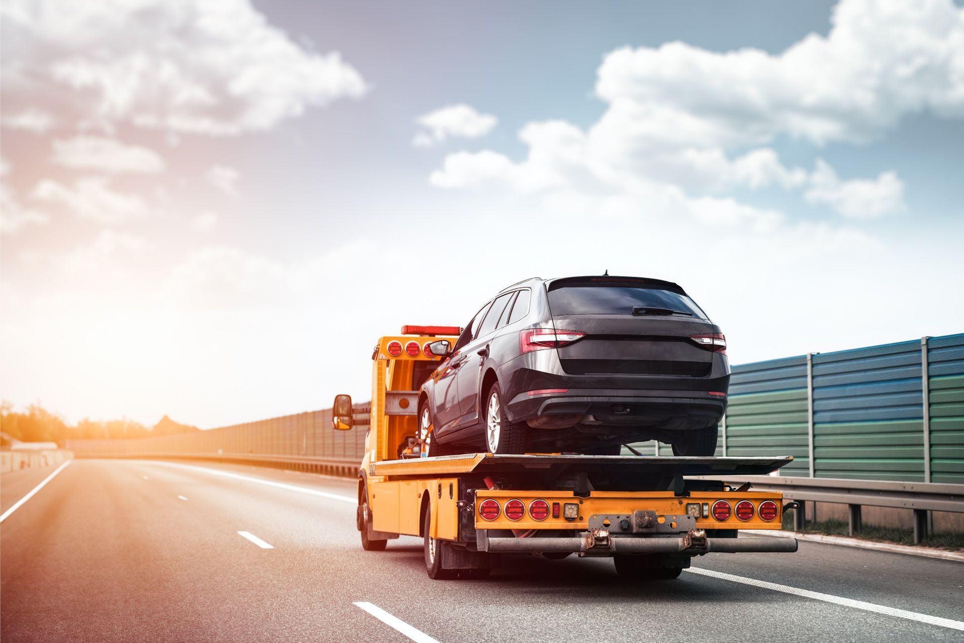 A black car loaded on a yellow tow truck driving on a highway on a sunny day.