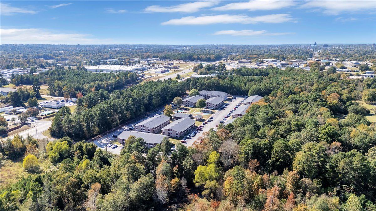 Aerial view of apartment complex surrounded by trees, with a town in the distance under a blue sky.