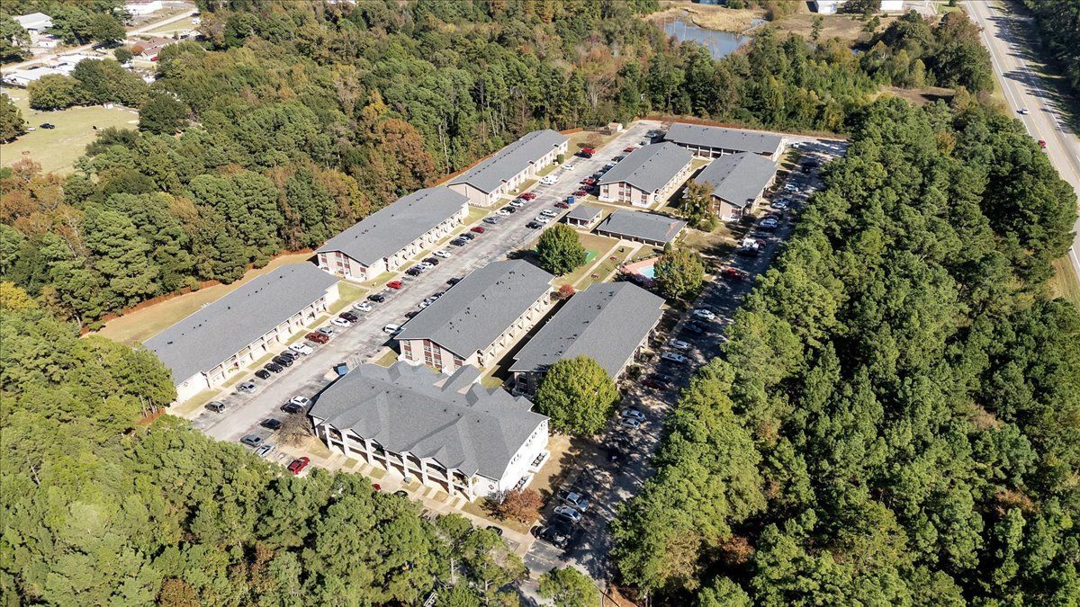 Aerial view of apartment complex surrounded by trees, with a road and a small pond visible.