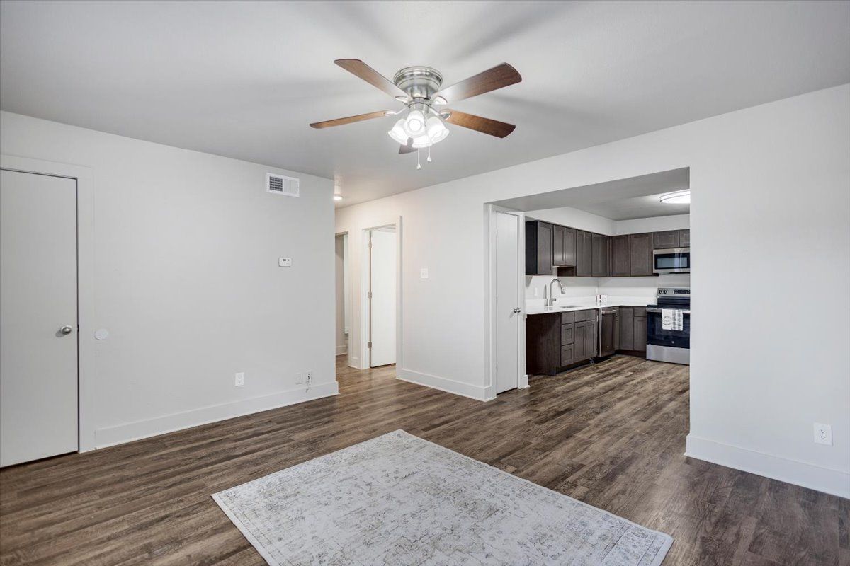 Empty apartment living room with brown wood flooring, light walls, and a view into the kitchen.
