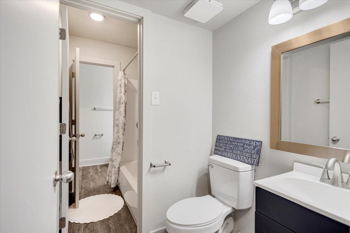 Bathroom with a toilet, sink, mirror, and shower, with wood-look flooring and white walls.