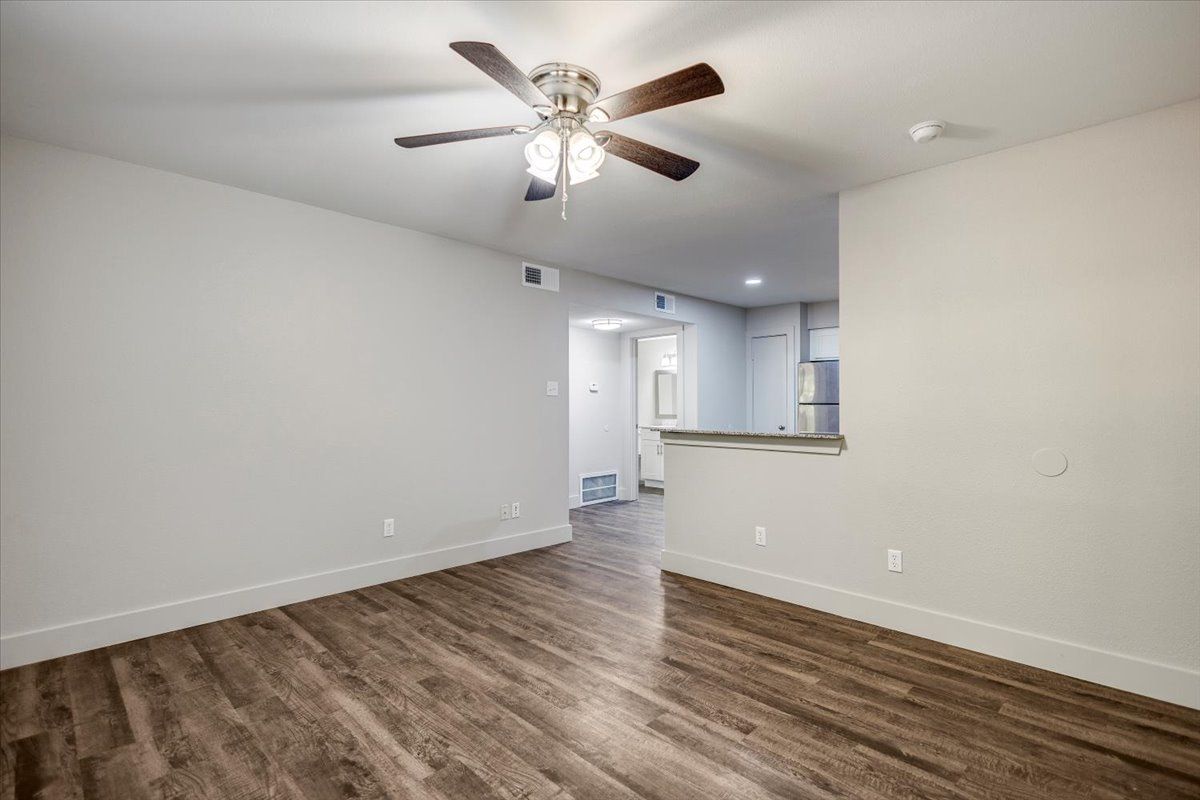 Empty room with wood-look flooring, light gray walls, ceiling fan, and opening to a kitchen area.