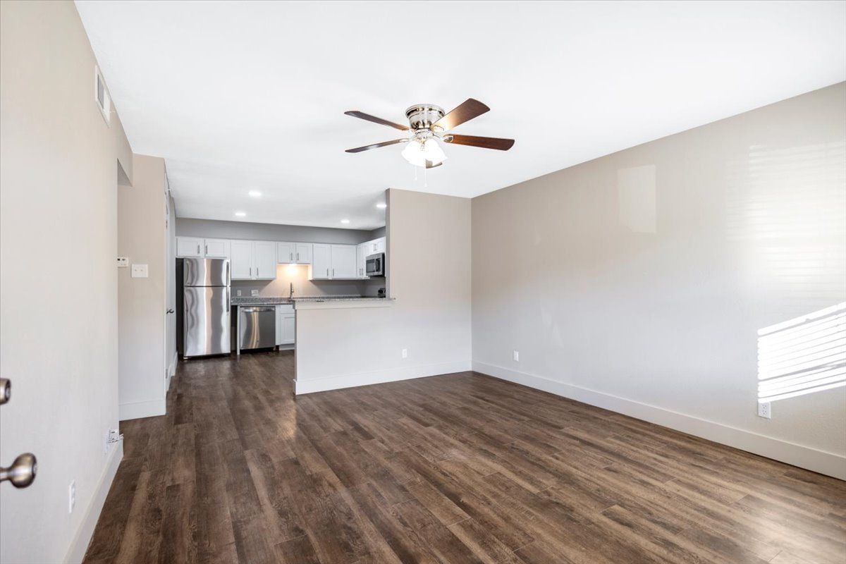 Empty living room with wood floors, neutral walls, and open kitchen visible in the background.