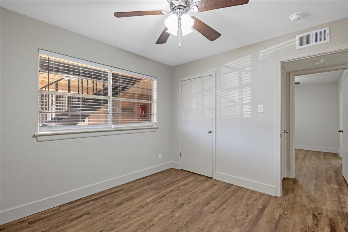 Empty bedroom with wood-look floors, white trim, a ceiling fan, and a window looking onto an exterior walkway.