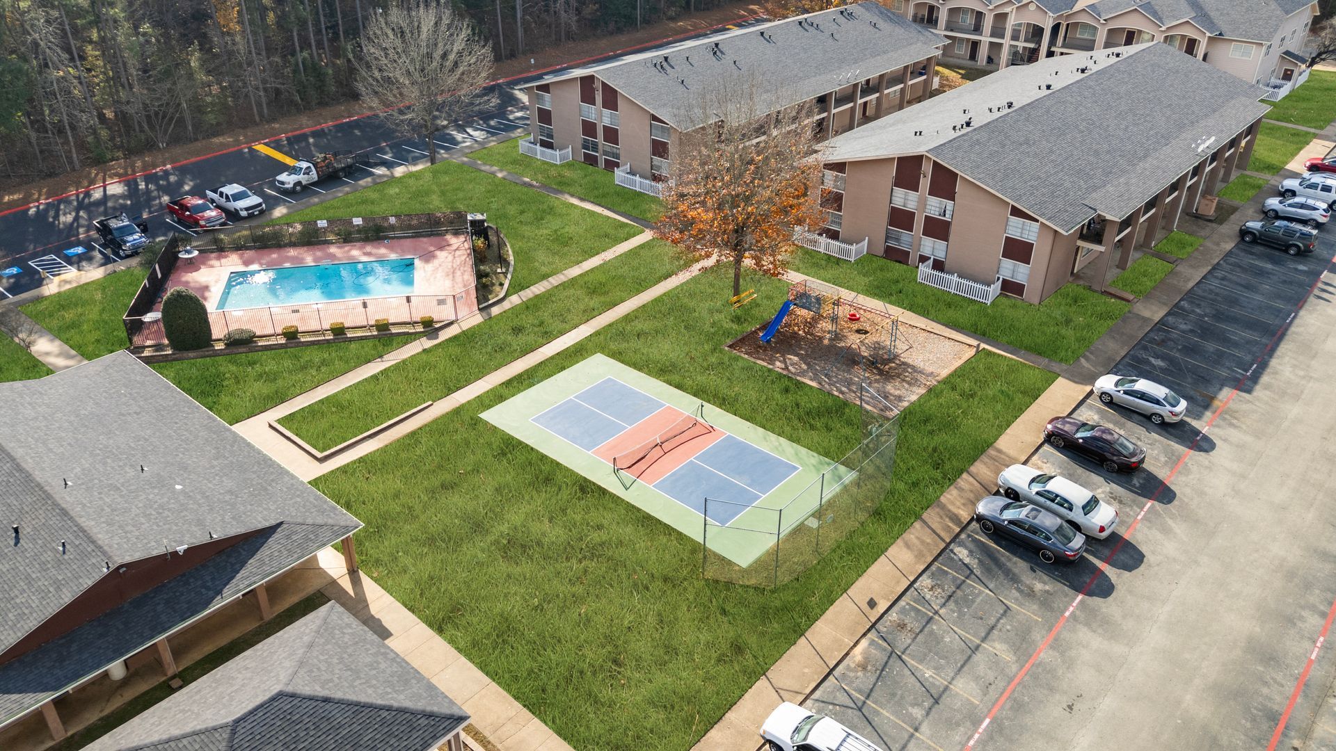 Exterior view of a two-story building complex with a sports court and cars in a parking area, under a blue sky.