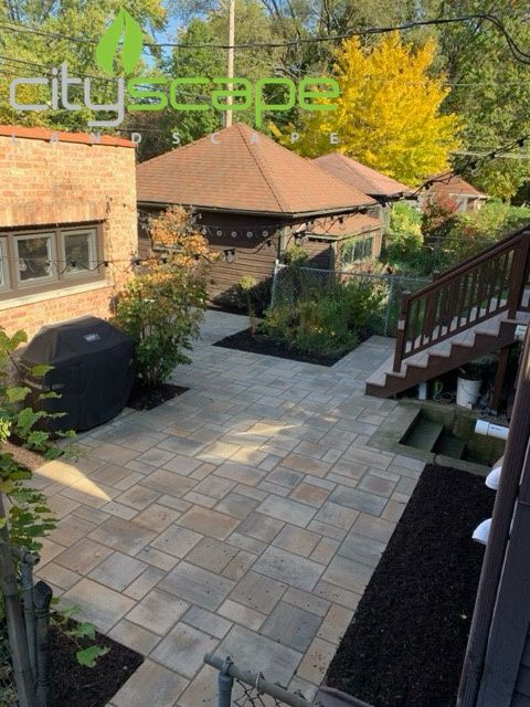 Backyard patio with brick pavers, raised deck, and small shed, sunny day.