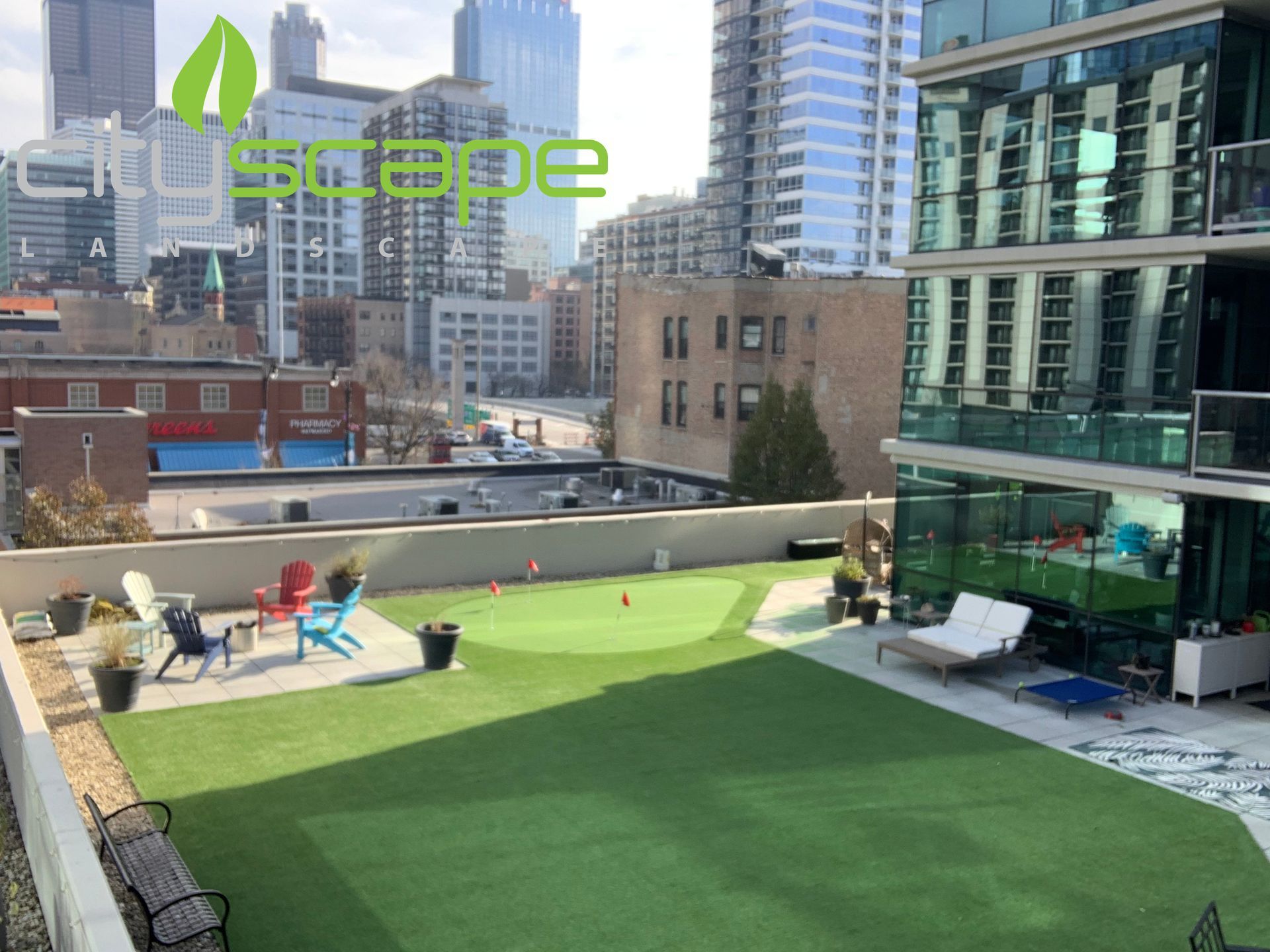 Rooftop putting green with city skyline in the background. Green artificial turf, lounge chairs, and potted plants.