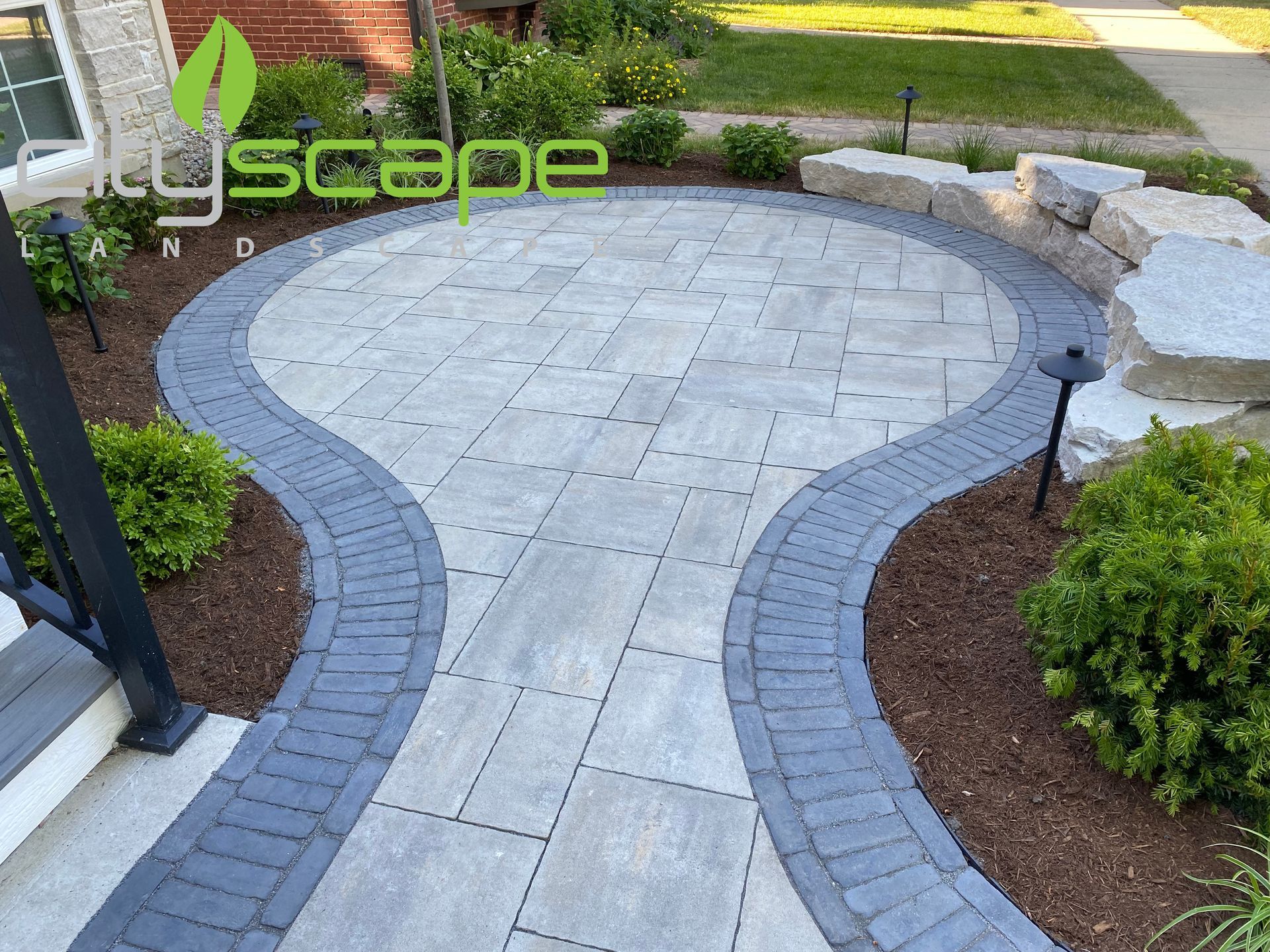 Stone patio and walkway with curved dark border, landscaping, and bushes.