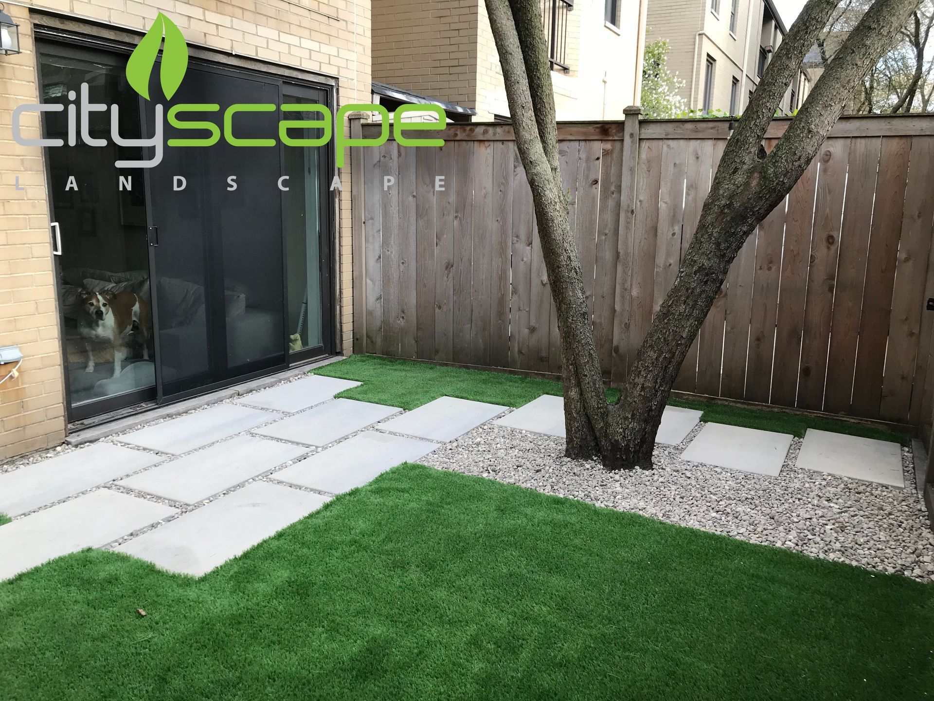 Backyard with artificial turf, stone pathway, wooden fence, tree, and sliding glass door.