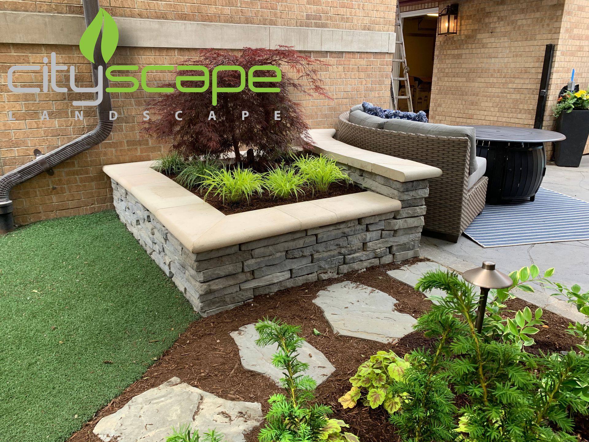 Stone and metal gate entrance, flanked by gabion and rust-colored metal walls. Gray sidewalk, blue sky.