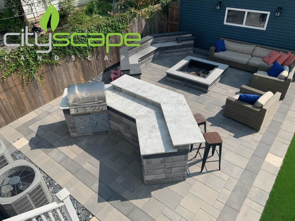 Outdoor patio with grill, bar, seating, and fire pit. Grey pavers and blue house. 