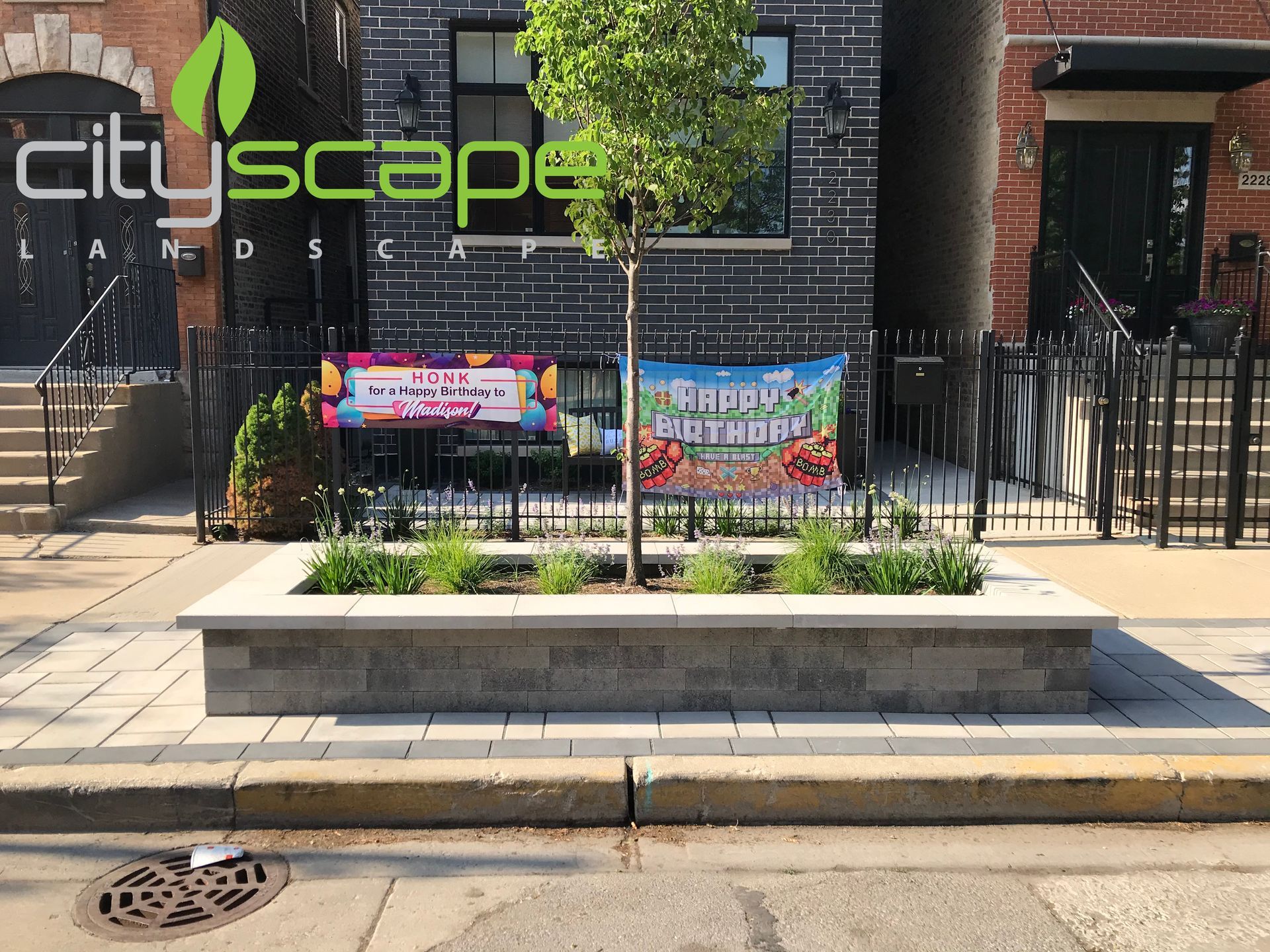 Streetscape with a tree and garden bed, in front of a building with promotional signs.