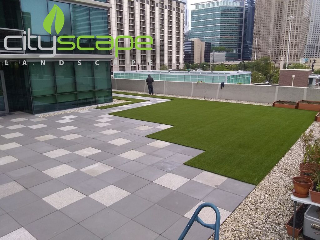 Rooftop with tiled patio, artificial turf, and a cityscape backdrop. A person walks near the center.