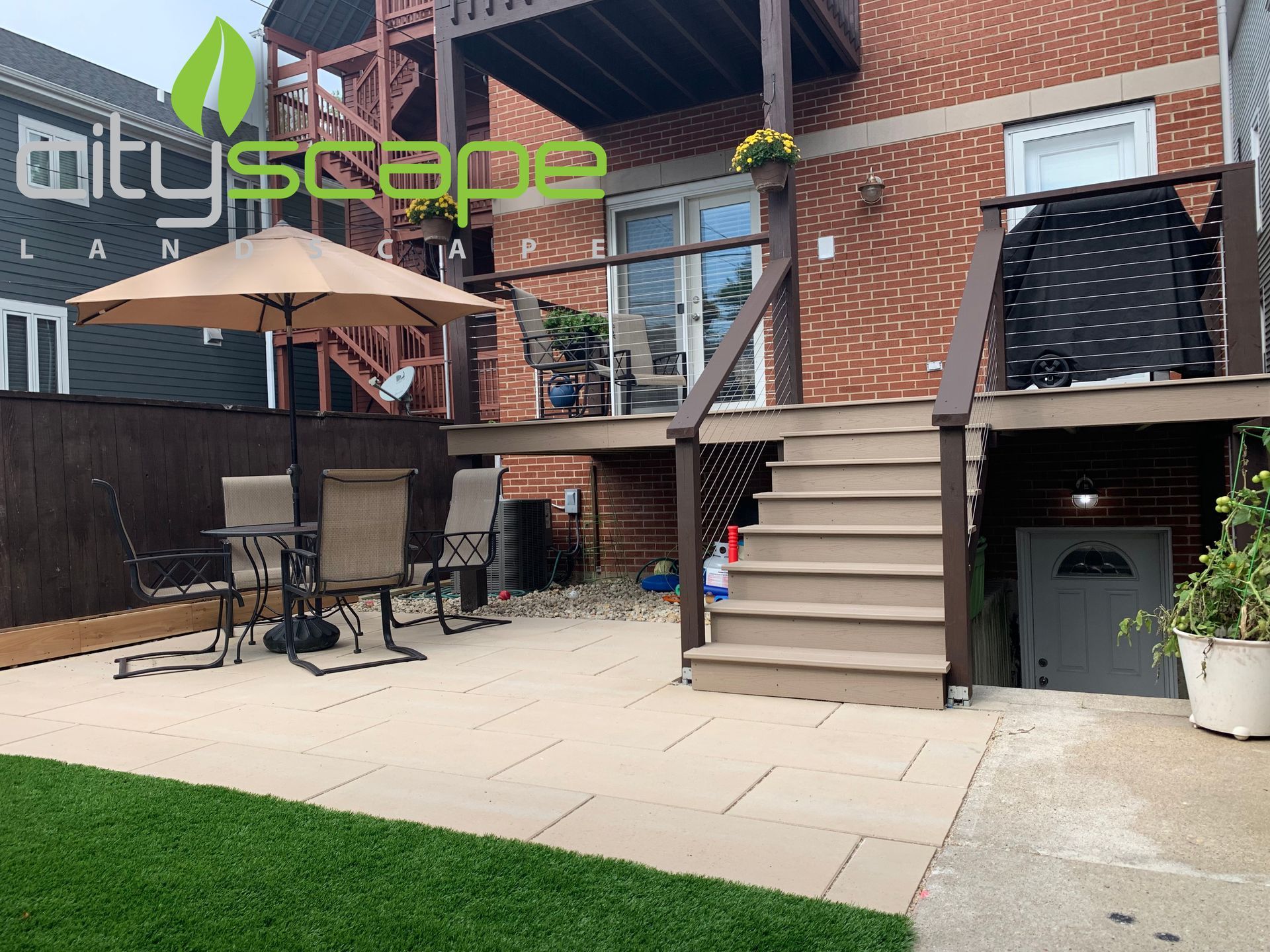 Backyard patio with steps up to a deck and brown umbrella, surrounded by grass and a brick building.