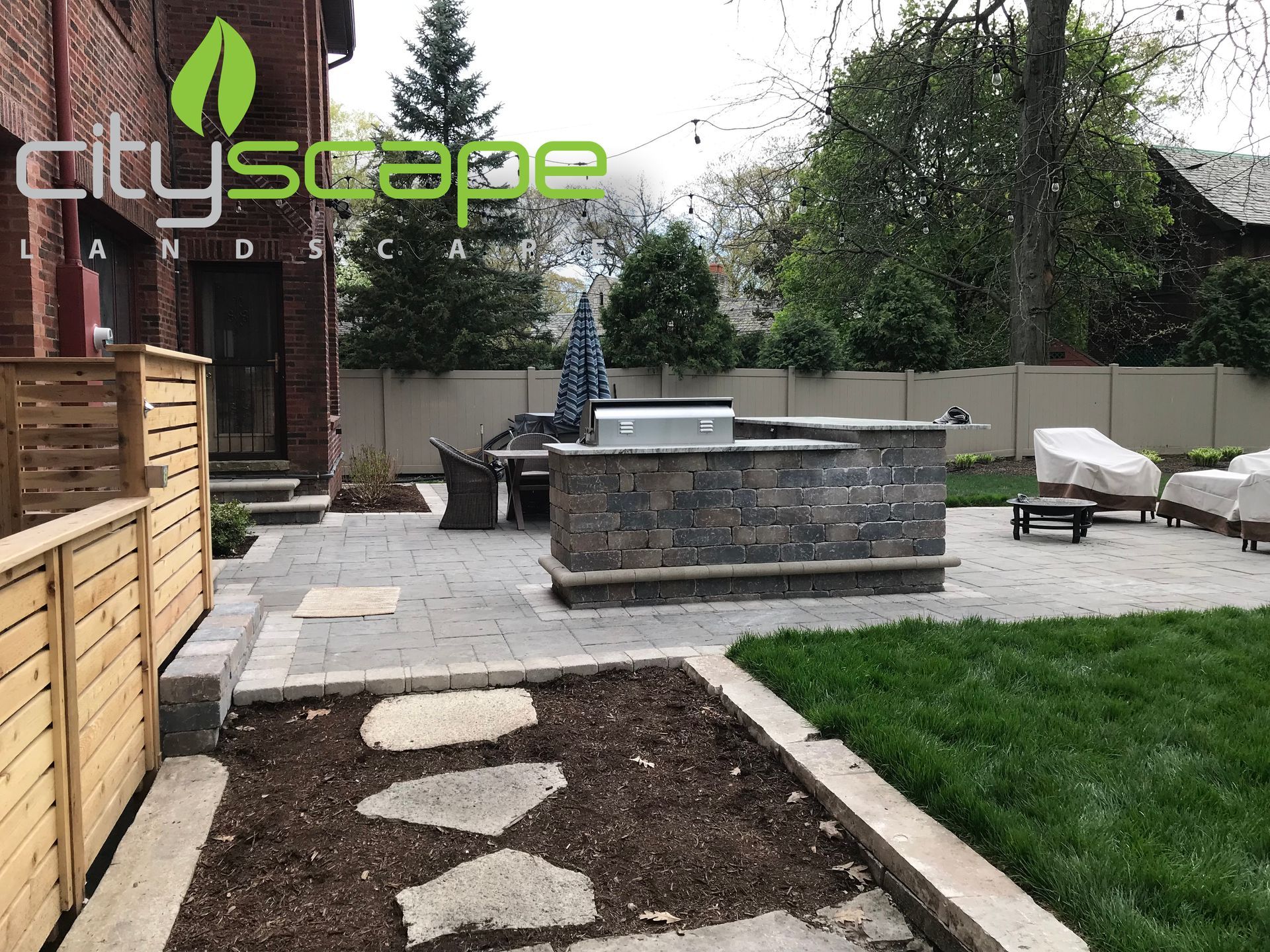 Backyard patio with stone grill, seating area, wooden fence, and green lawn.