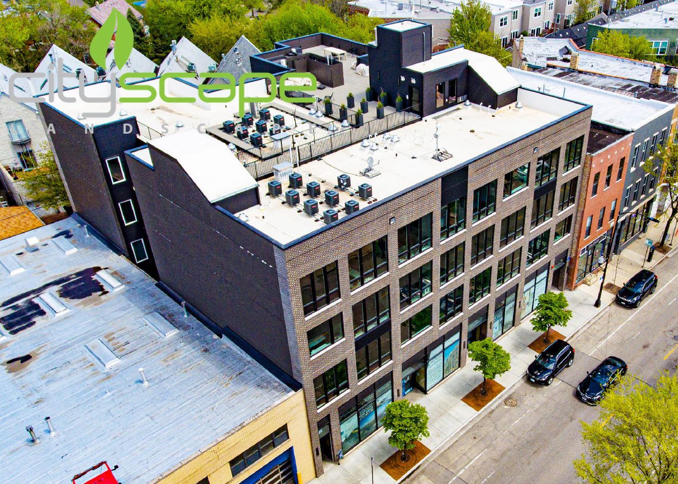 Aerial view of a modern brick building with large windows and a flat roof, street level with trees and parked cars.