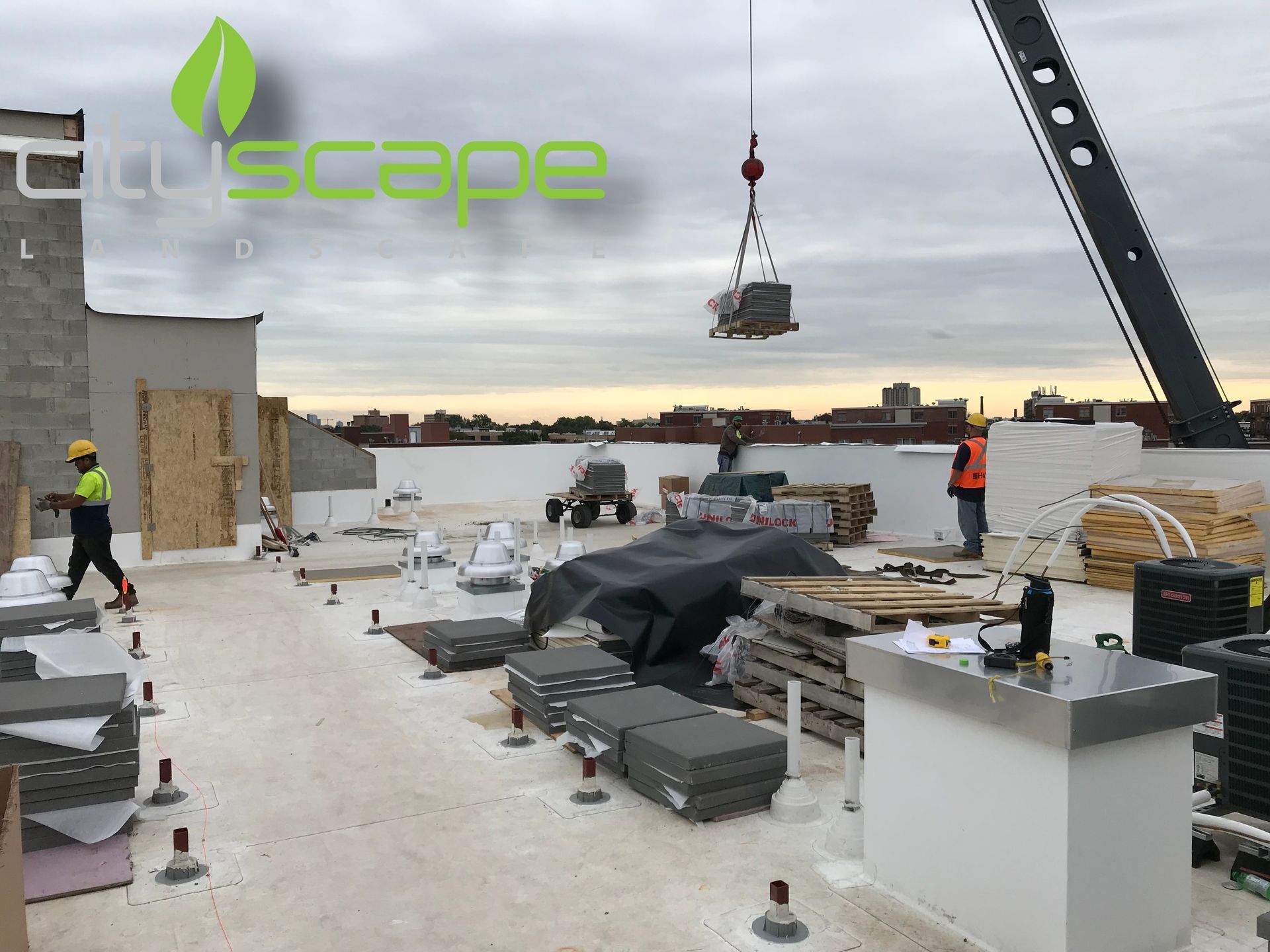Rooftop construction site with crane lifting equipment. Workers in safety gear, white roof, cloudy sky.