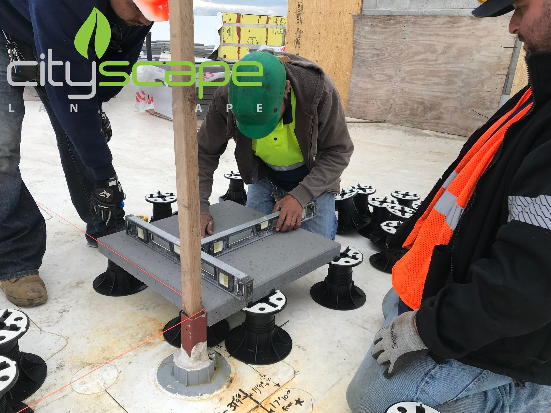 Construction workers using a level to install a grey paver supported by adjustable pedestals on a rooftop.