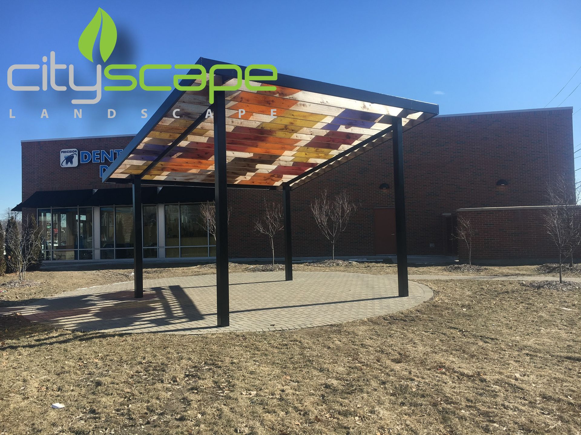 A black metal pergola with a colorful stained-glass roof sits on a brick patio in front of a brick building.