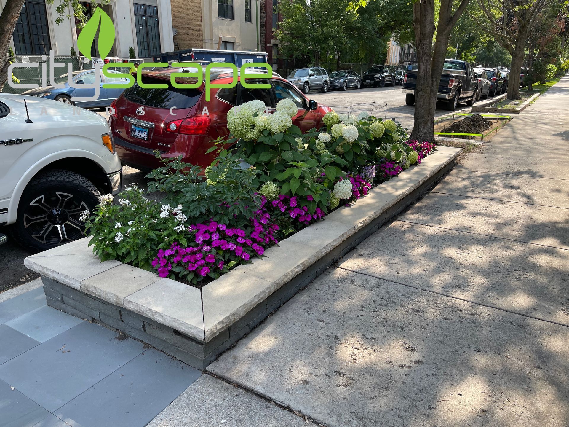 Stone planter box with colorful flowers next to a sidewalk and parked cars.