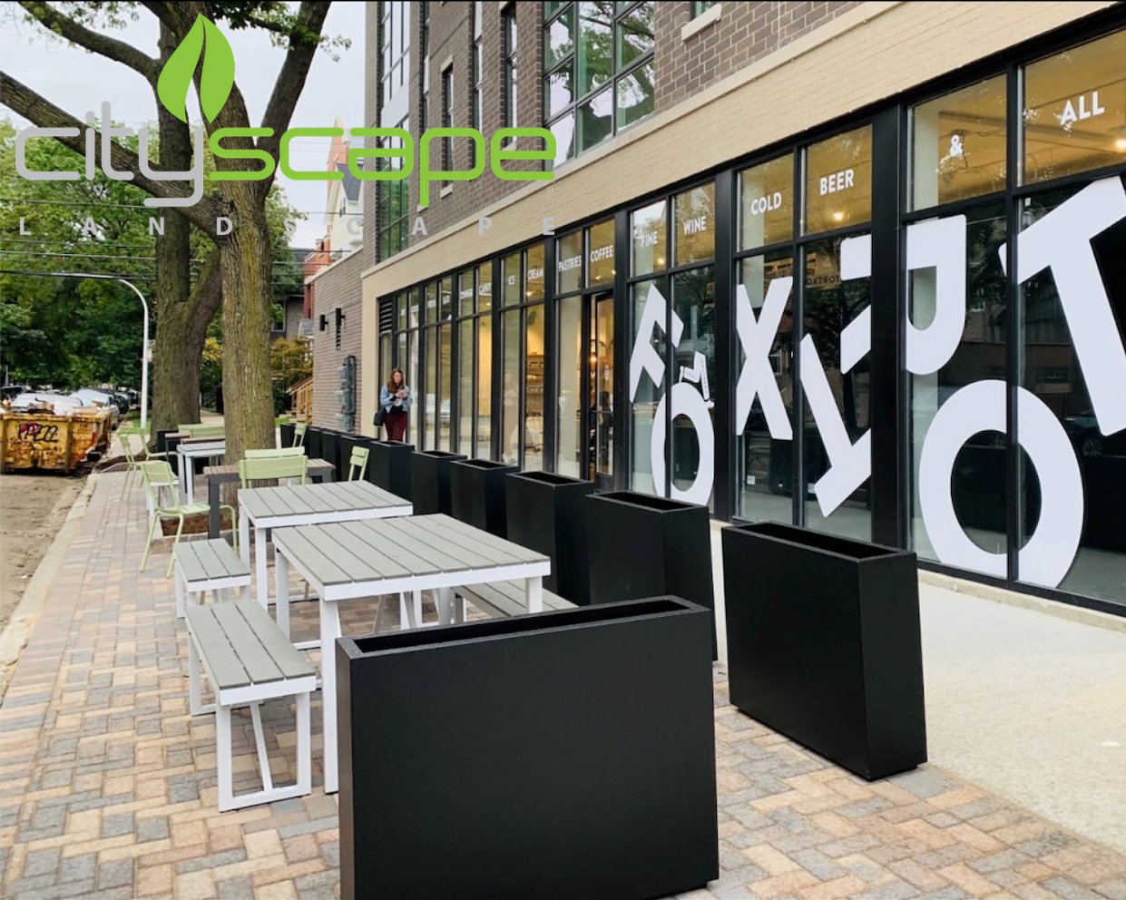 Outdoor seating area with tables and benches, flanked by large black planters, in front of a building with large windows.
