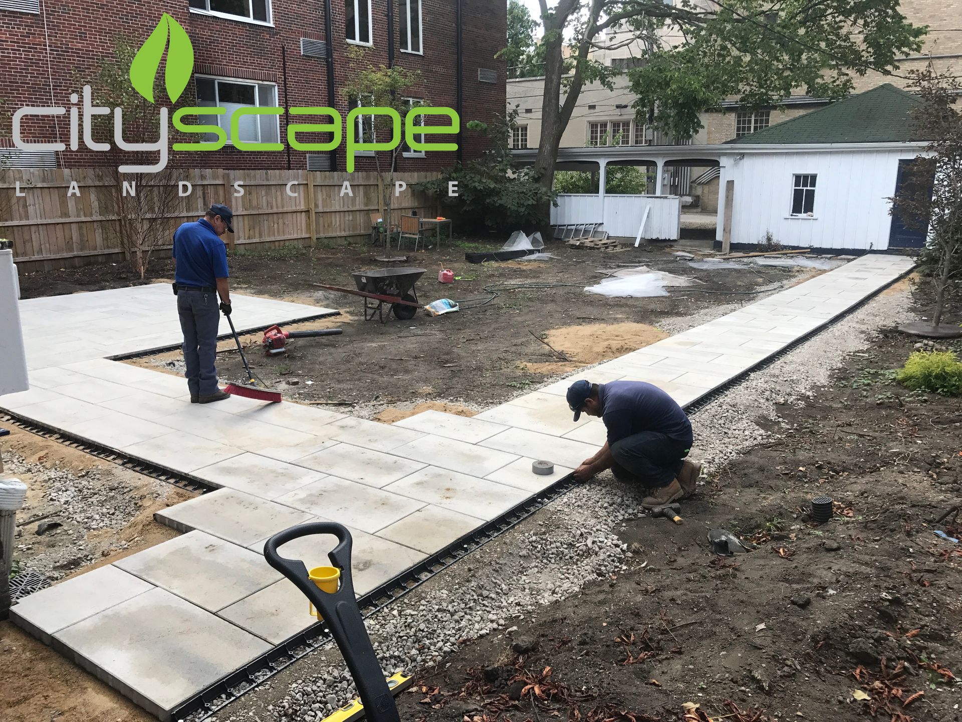 Two workers laying pavers for a patio in a backyard; one sweeps, the other kneels and adjusts.