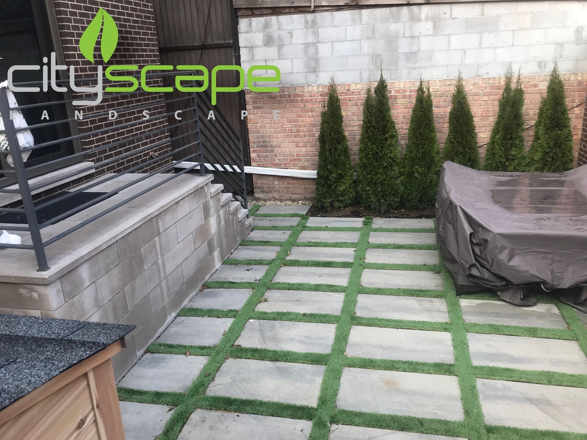 Patio with square pavers and artificial grass, featuring a covered table and evergreen trees.