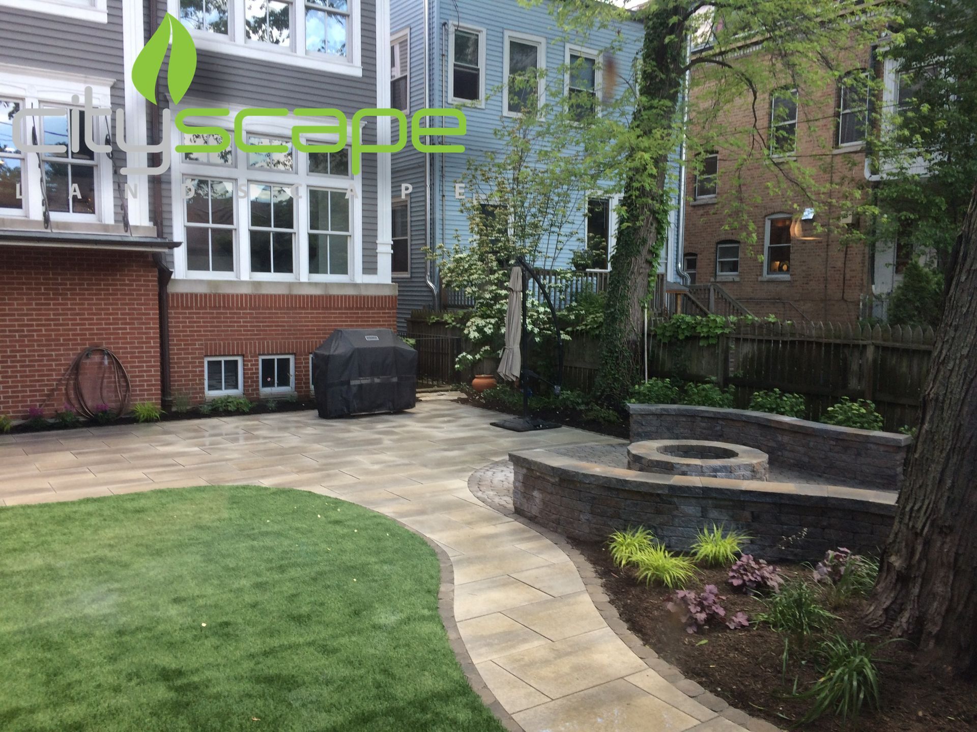 Backyard patio with paved path, grass, stone wall, fire pit, and grill.