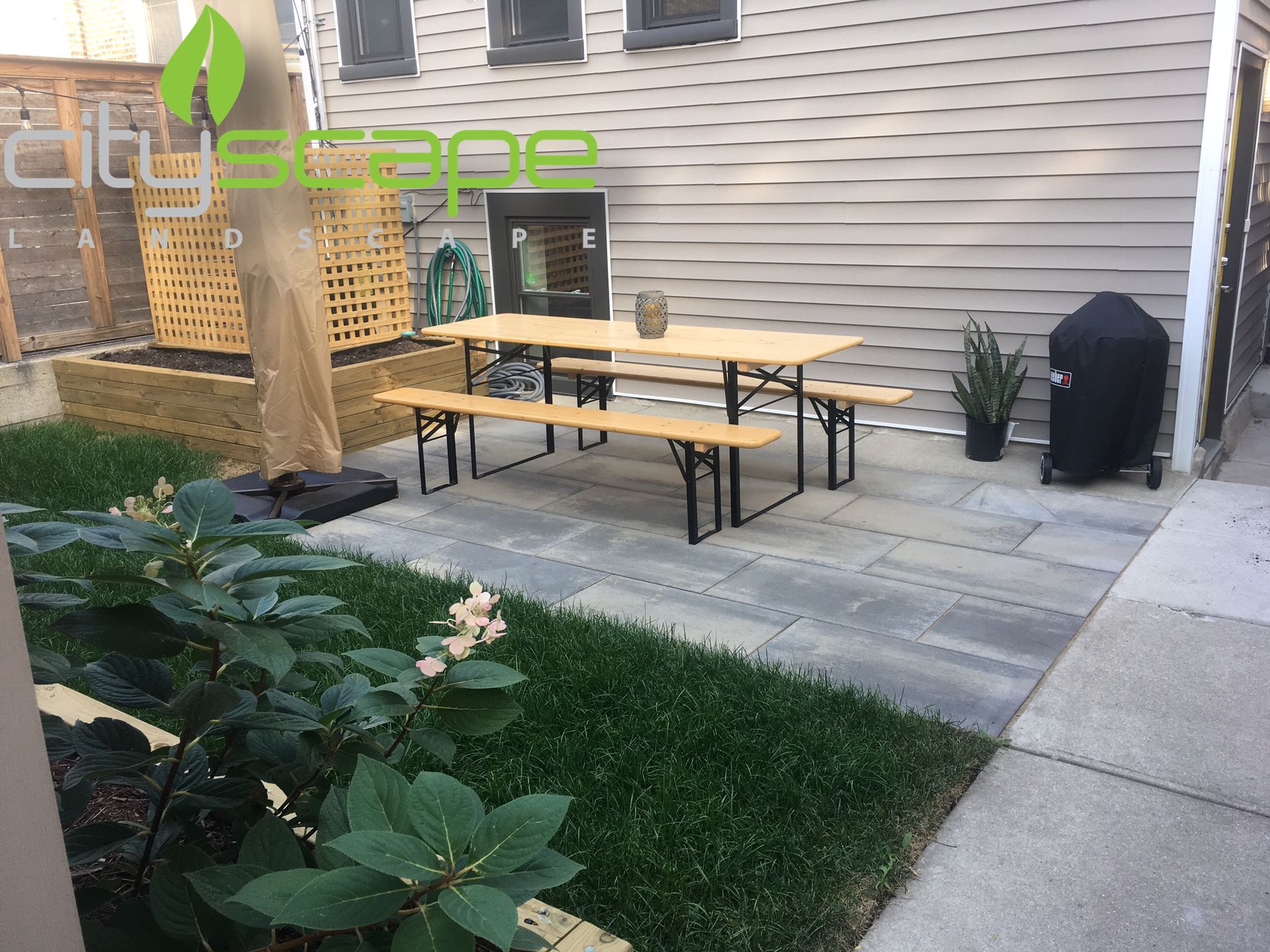 Backyard patio with wooden picnic table, grill, gray pavers, greenery, and a lattice fence.