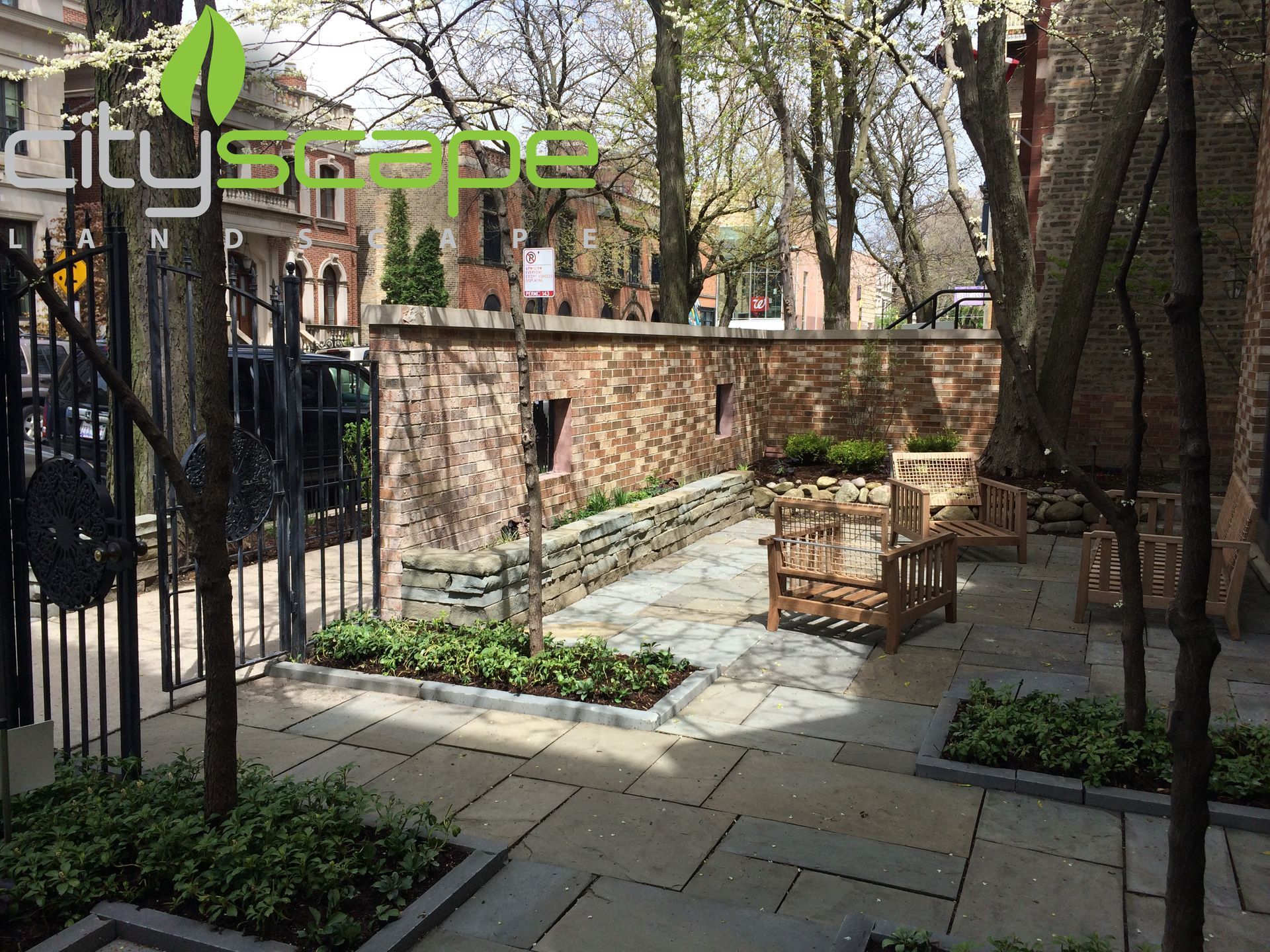 A small courtyard with brick walls, stone paving, seating, and landscaping; trees frame a view of buildings.
