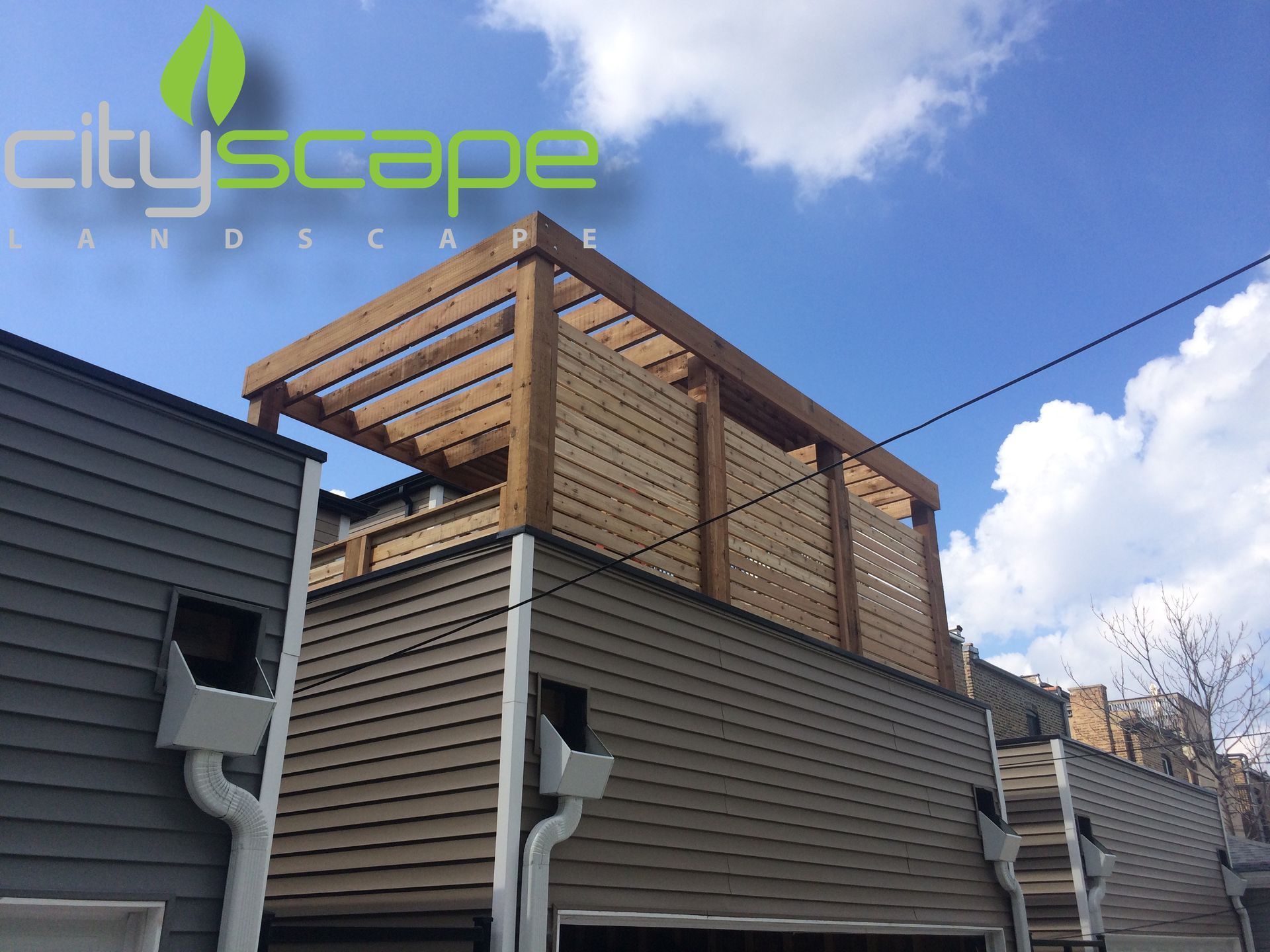 Wooden rooftop pergola with lattice screen, brown siding, against a blue sky with clouds.