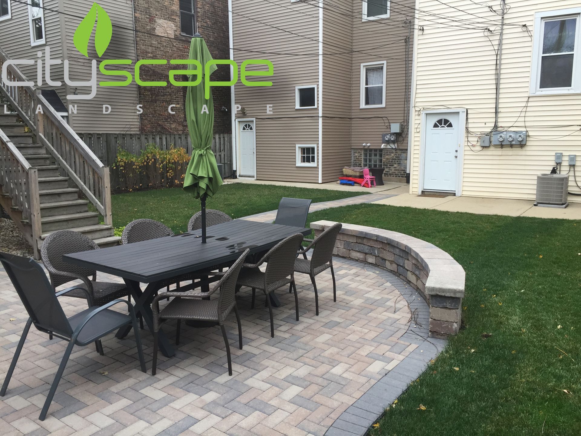 Backyard patio with table and chairs on pavers; green umbrella, grass, and building in the background.