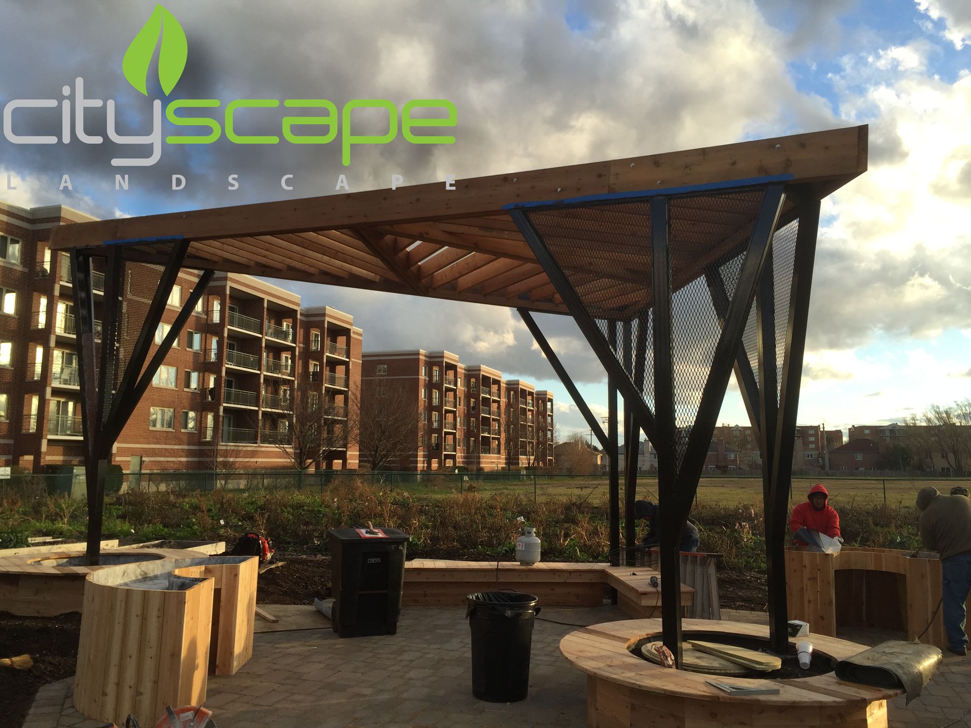Wooden and metal pergola with seating, in front of a modern apartment building.