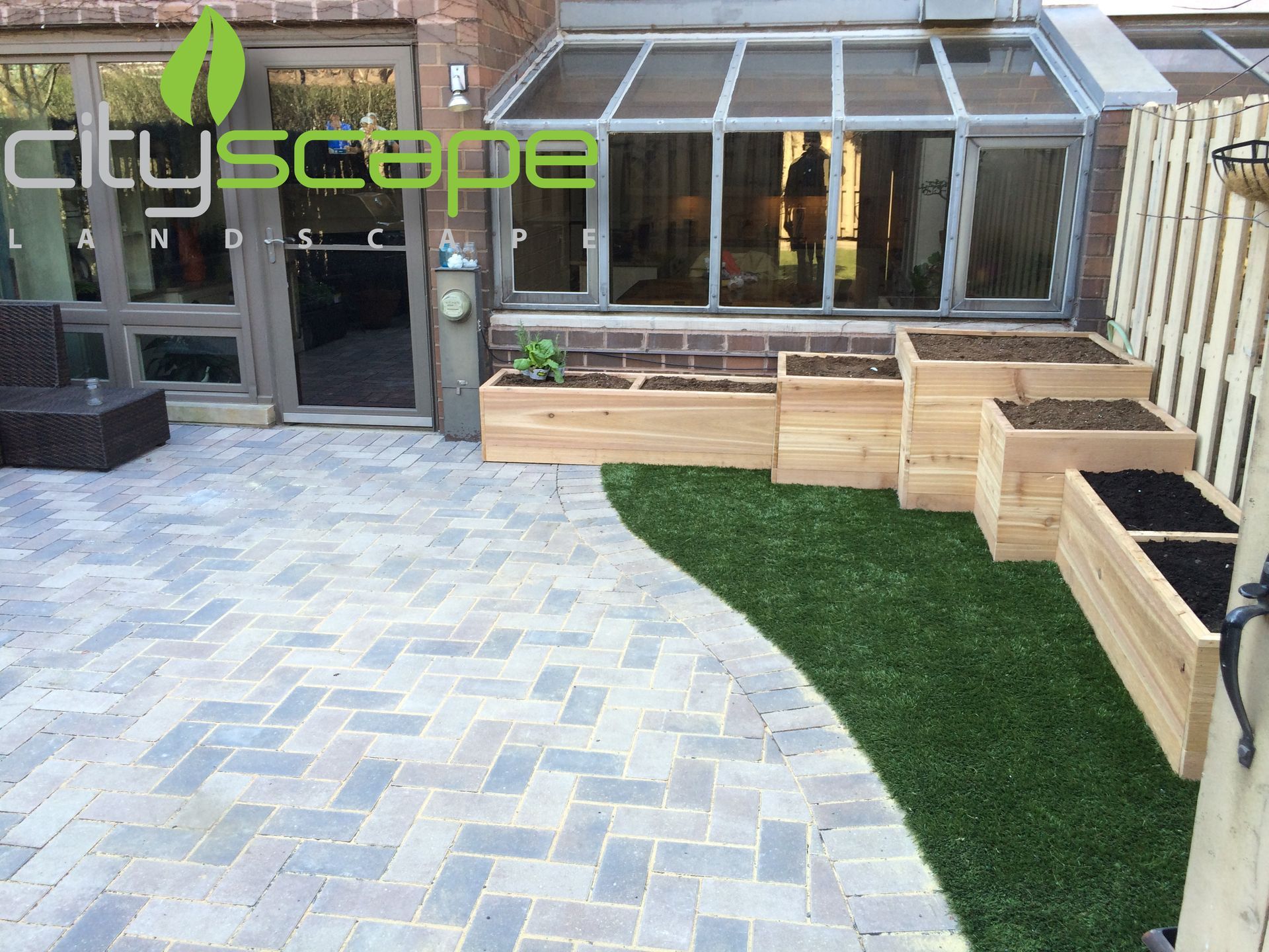Patio with brick pavers, raised wooden planters, and a strip of green turf.