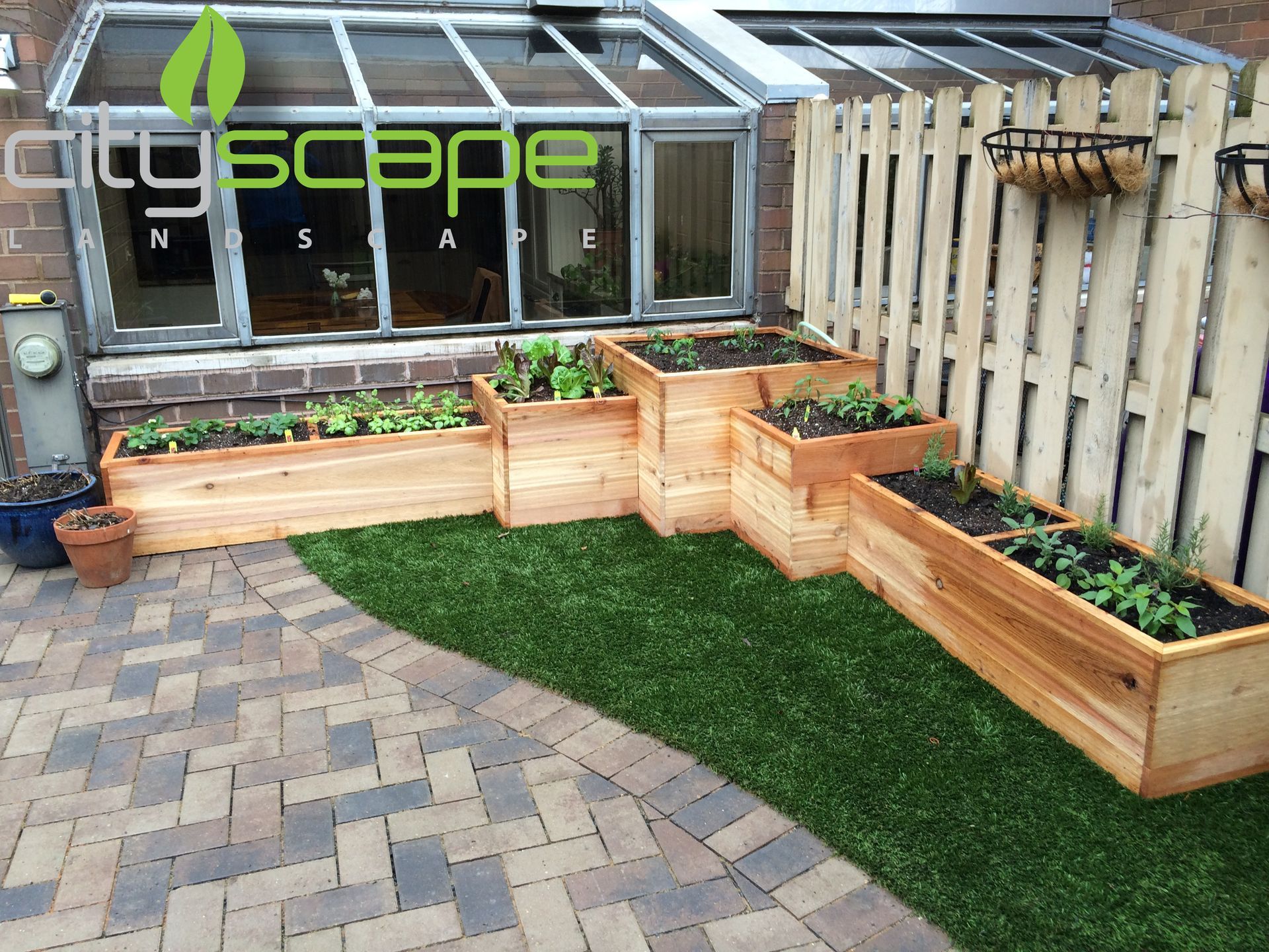 Raised wooden garden beds with plants sit on artificial turf beside a brick patio and a fence.