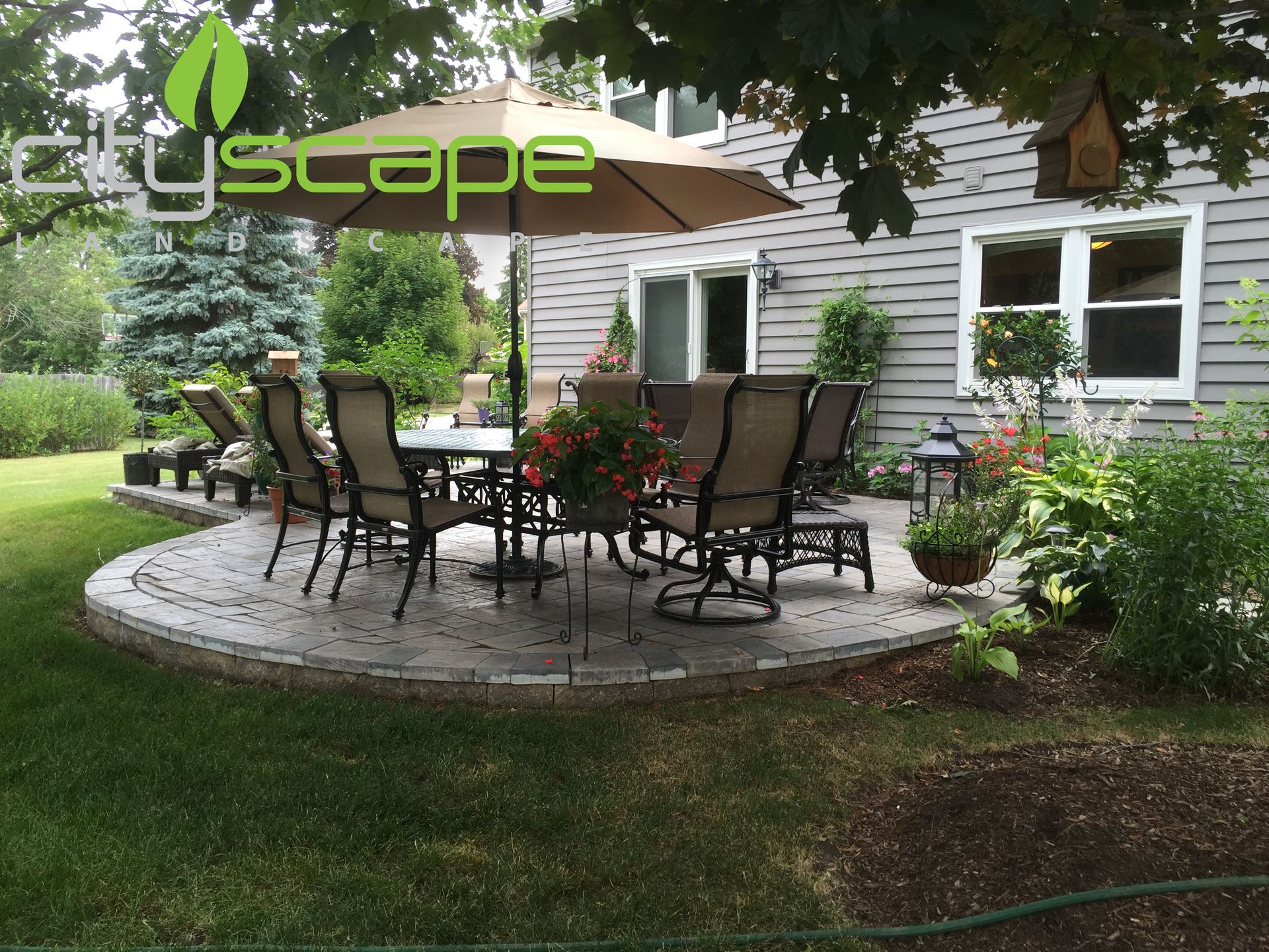 Patio with dining set, umbrella, and surrounding landscaping, next to a house.
