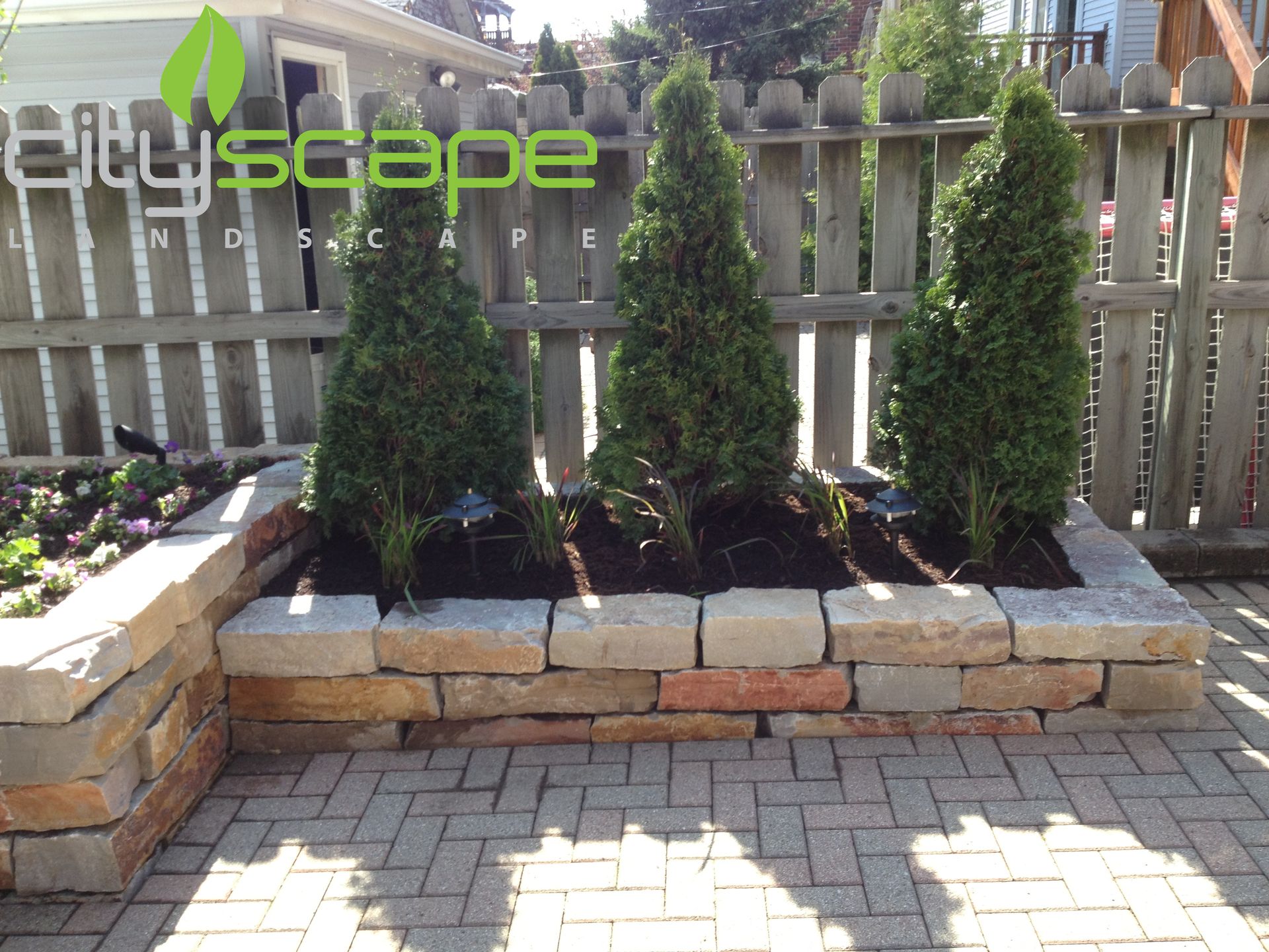 Raised garden bed with three evergreen trees against a wooden fence. Brick pavers and stone blocks.