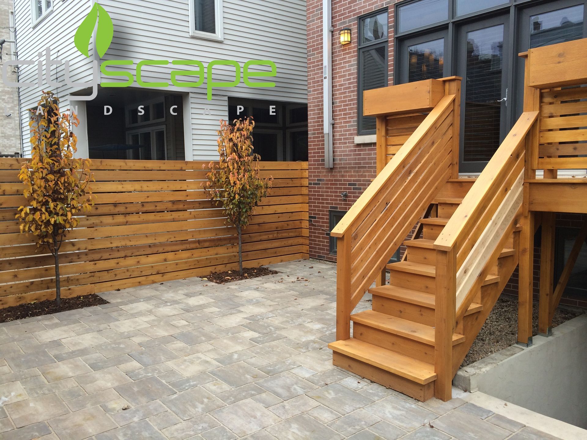 Wooden deck and stairs leading to a patio with a horizontal slat fence, two small trees, and brick building.