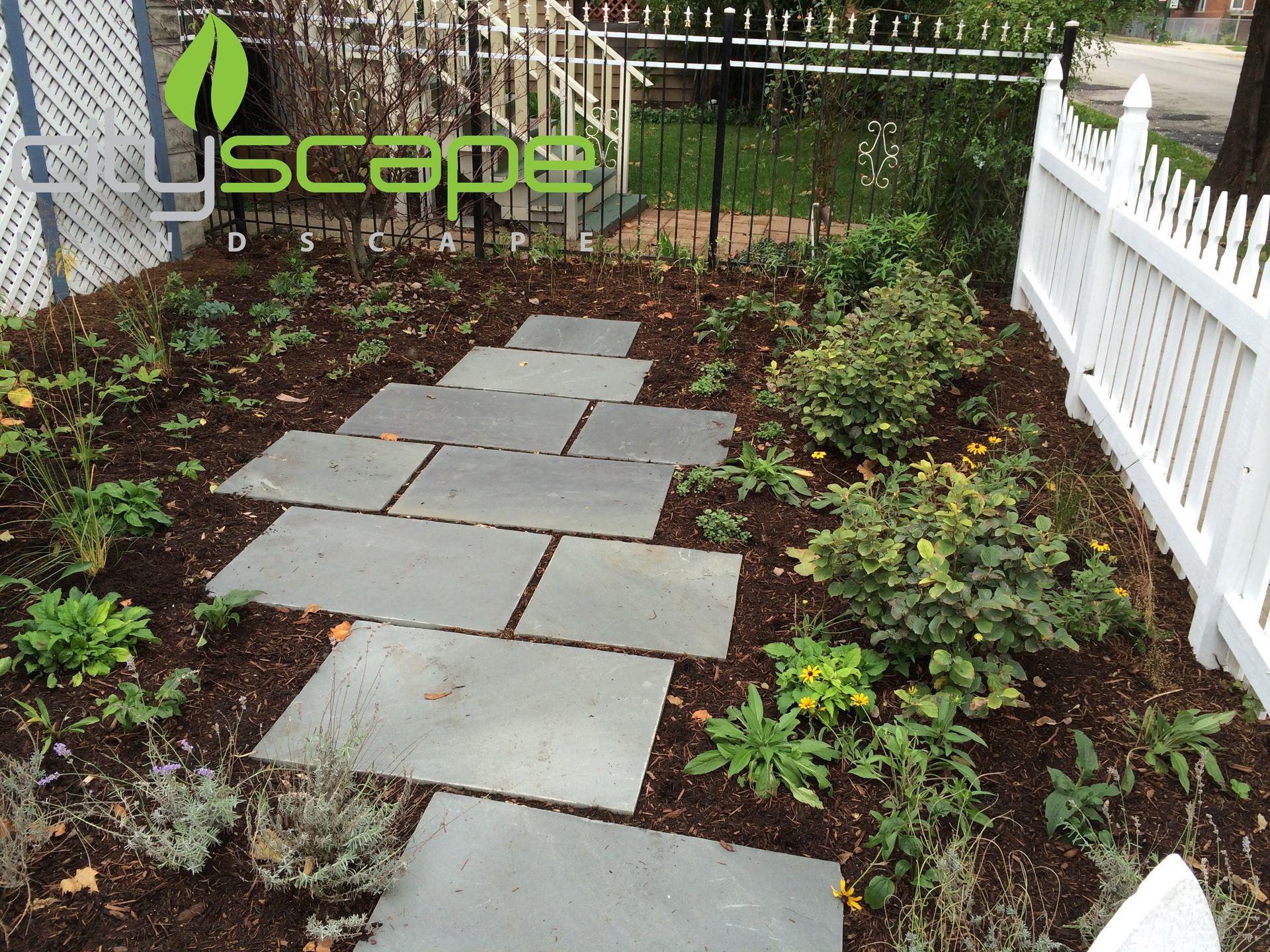 Stone pathway through a garden with a white picket fence.