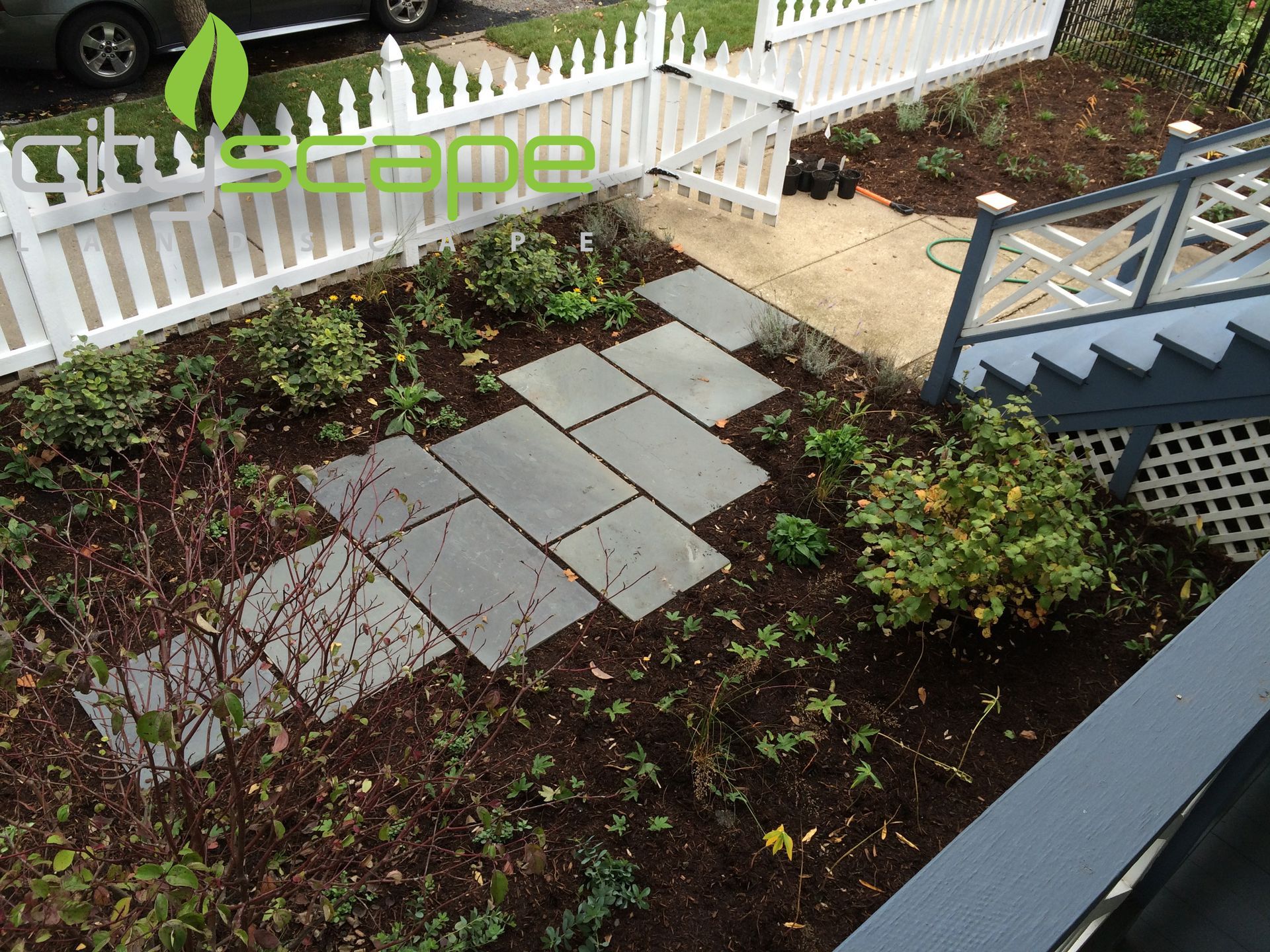 Stone pathway through a garden bed with mulch, plants, and a white picket fence.