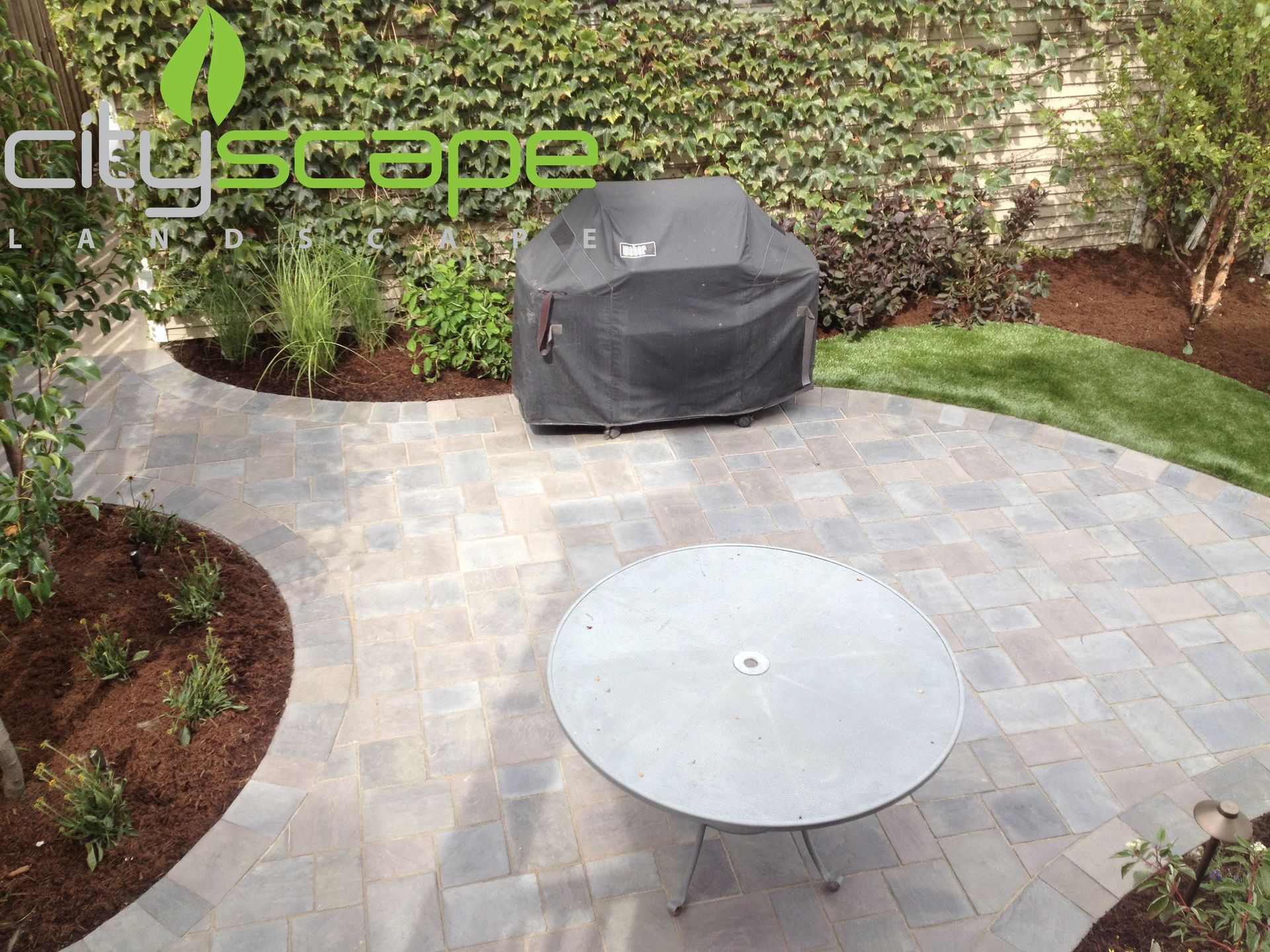 Patio with paving stones, table, covered grill, and landscaping.