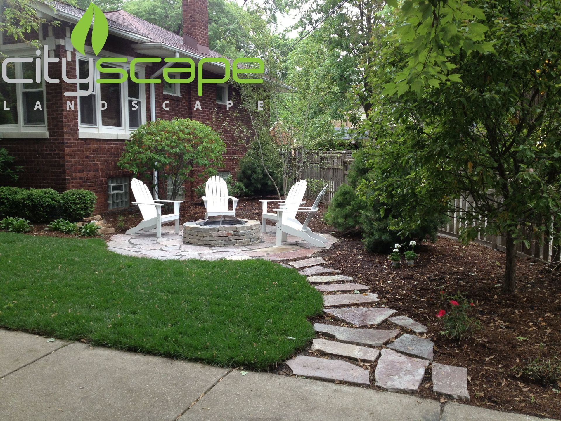 A backyard with fire pit, white chairs, stone pathway, brick house, and green lawn.