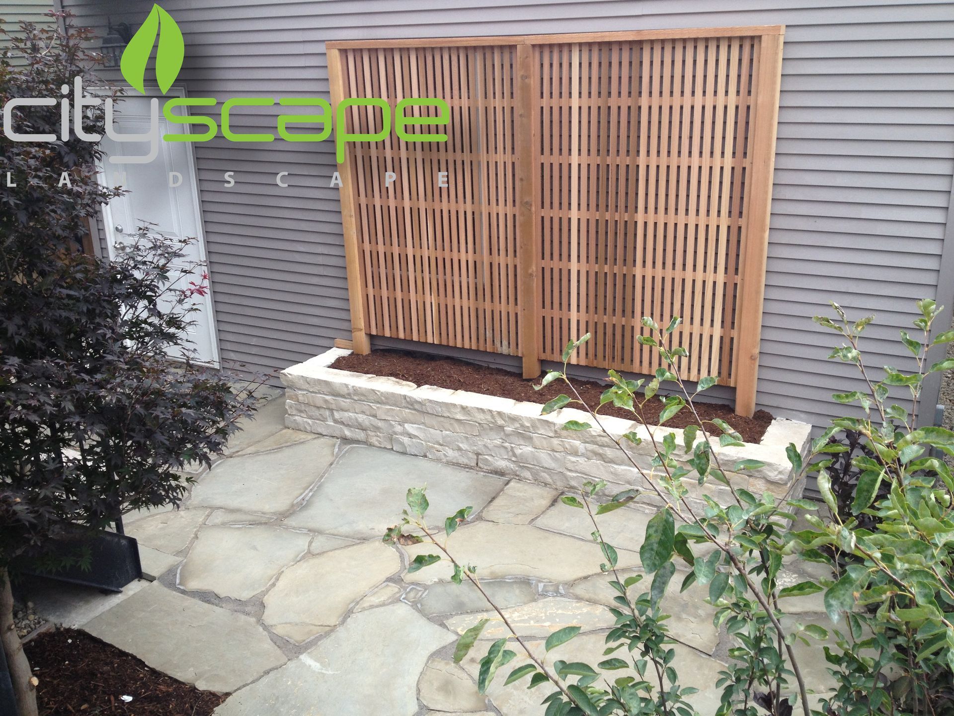 Stone path leads to a wooden privacy screen on a raised stone planter, with landscaping.
