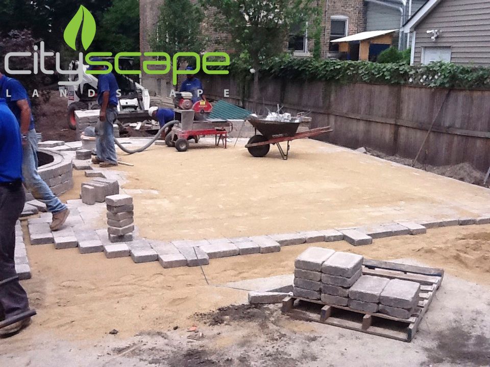 Workers laying pavers in a yard, with stacks of bricks, sand, and tools present.