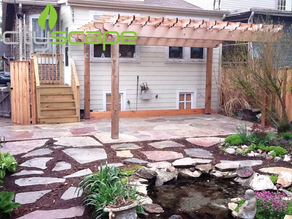 Backyard patio with stone path, pergola, pond, and wooden stairs against a light-colored house.